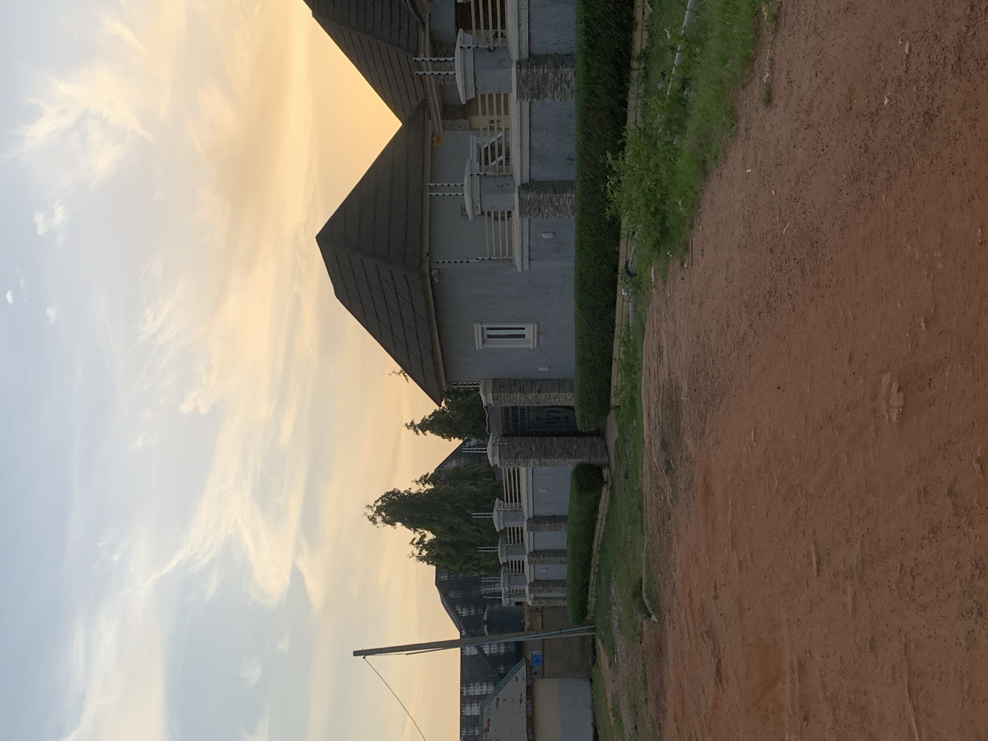 The image captures a street view in Rido, Nigeria, at dusk. The foreground is dominated by a dirt road. To the right, a neatly manicured hedge runs along a modern fence with stone pillars and a metal gate, enclosing two houses. The houses have light gray stucco exteriors and dark, conical roofs. Electrical fencing is visible along the top of the wall. In the background, a cluster of tall, slender trees stands behind the fence, with other buildings and structures partially visible behind them, including one with a corrugated metal roof. The sky above is illuminated by the warm tones of a setting sun, with wispy clouds stretching across the horizon. The overall impression is one of residential development in an urban or semi-urban environment.