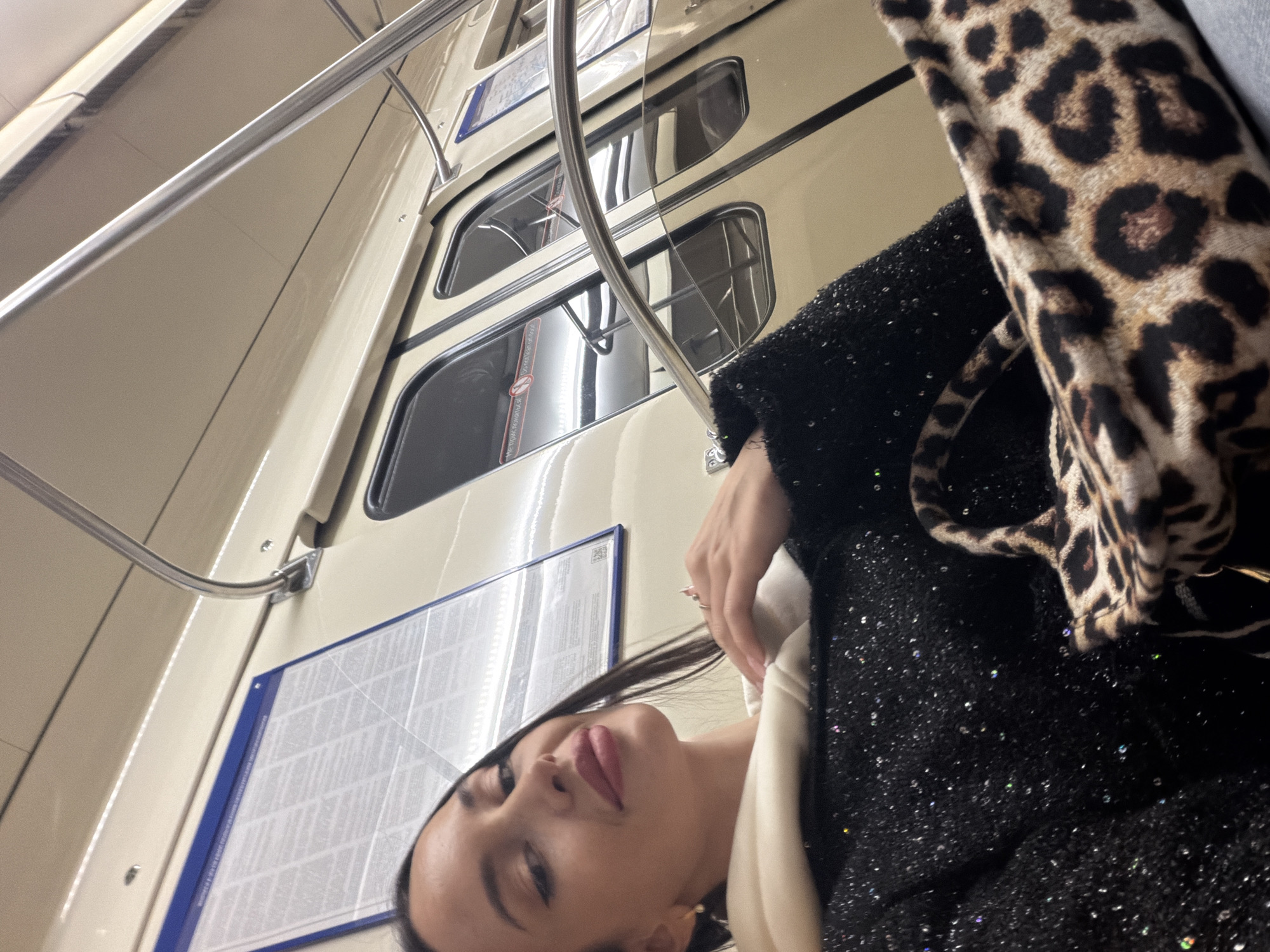The image shows a woman in a subway car. The subway car appears to be from the Saint Petersburg Metro, based on the design of the walls and windows. The woman is wearing a dark, possibly sequined, jacket and a light-colored scarf or top. She is also carrying a leopard-print bag. She is looking directly at the camera. The scene is lit with artificial light.