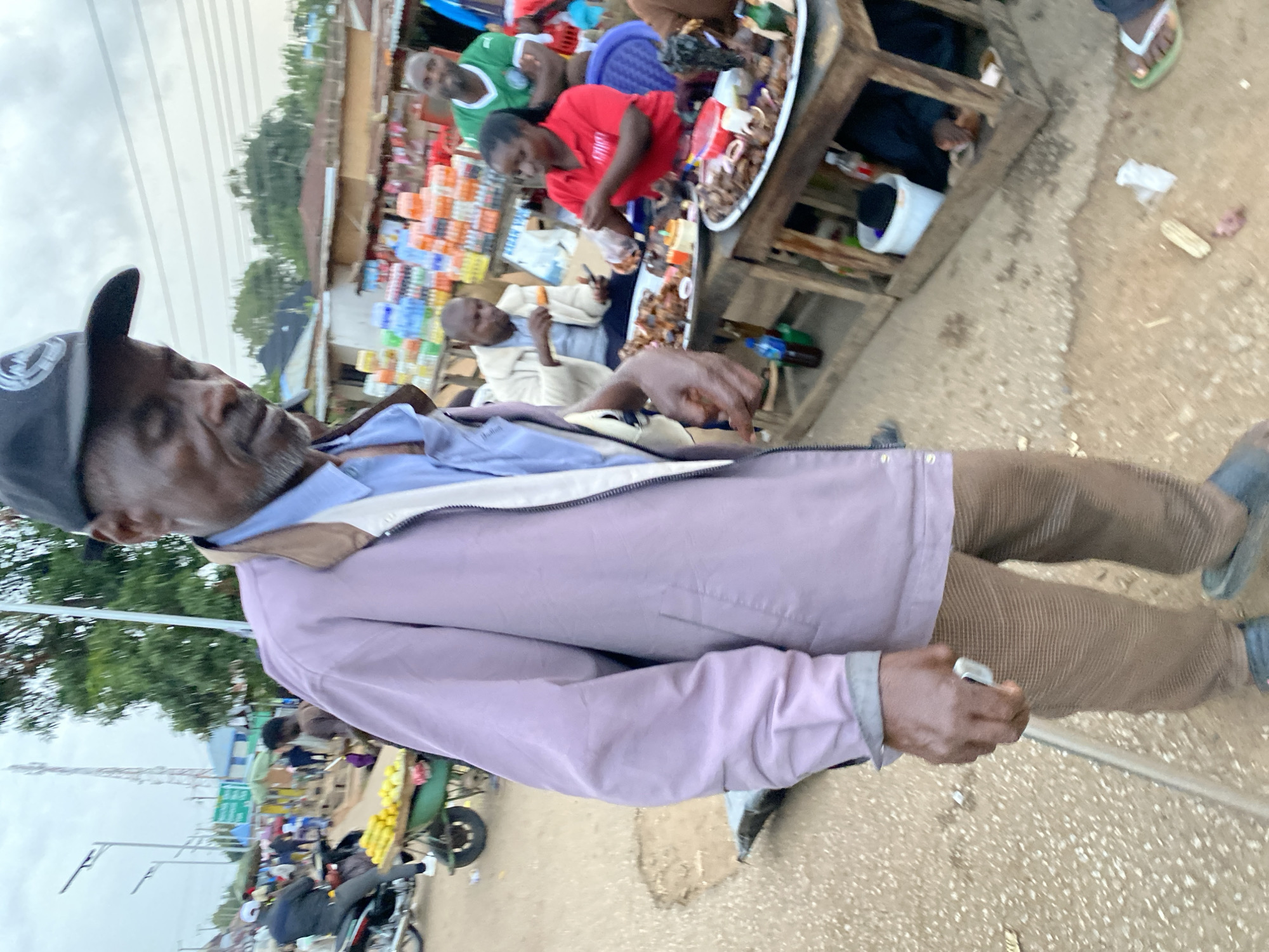 In the city of Bukuru, Nigeria, an elderly man with a cane, wearing a light purple jacket over a blue shirt and patterned trousers, stands in the foreground, slightly to the left. He is looking down and to his left. Behind him, a busy street scene unfolds. To the left, a vendor pushes a cart laden with yellow fruits, possibly lemons or mangoes, while a person on a motorcycle waits nearby. In the background, market stalls are visible, with rows of bottled drinks stacked on shelves. Several people are interacting at the stalls, some appearing to be vendors serving customers. One woman in a red shirt is handing food to a customer. Another man in a green shirt stands behind the woman. The ground is dirt and appears somewhat uneven, with scattered debris. The sky is overcast.