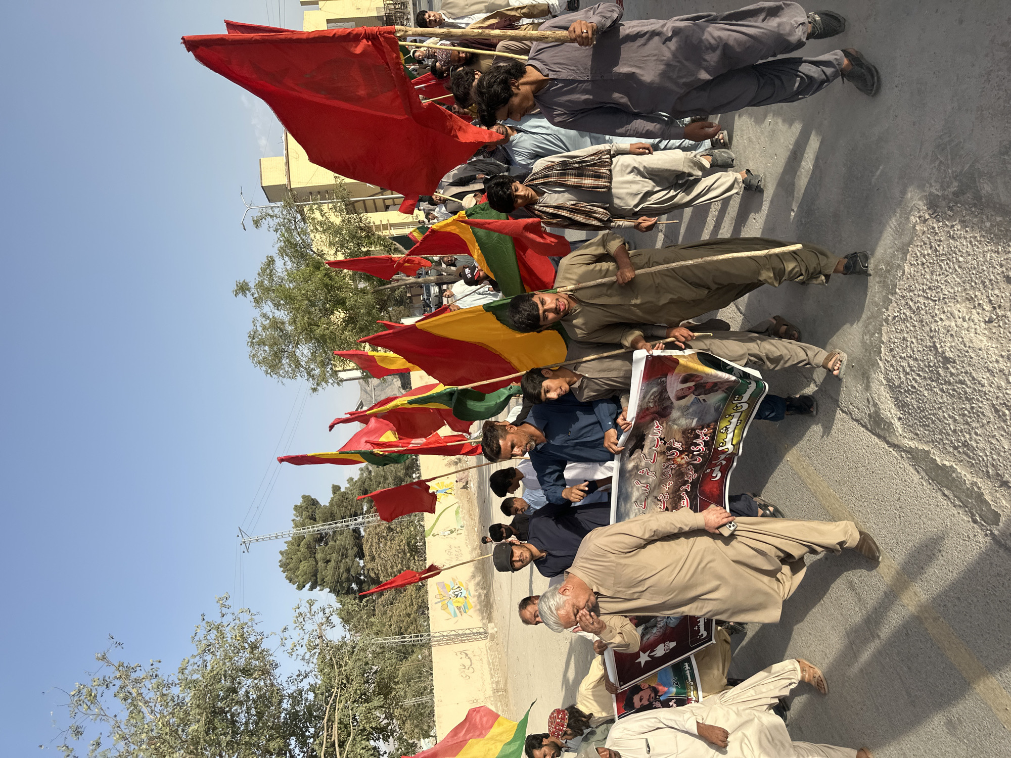 A group of men are marching outdoors, carrying flags and a banner. The men are wearing traditional Pakistani clothing, primarily shalwar kameez in various shades of beige, brown, and grey. Some are holding long wooden poles with flags attached, which are predominantly red, but also include yellow and green. The banner, held by several individuals in the foreground, has images and text in Urdu. The environment appears to be a street or road with a paved surface, bordered by a low wall with graffiti. Buildings and trees are visible in the background, under a clear blue sky. The lighting suggests it is daytime, with shadows indicating the sun's position. The men appear to be participating in a procession or demonstration, given the flags and banner. This scene is located in Quetta City Tehsil, Pakistan.