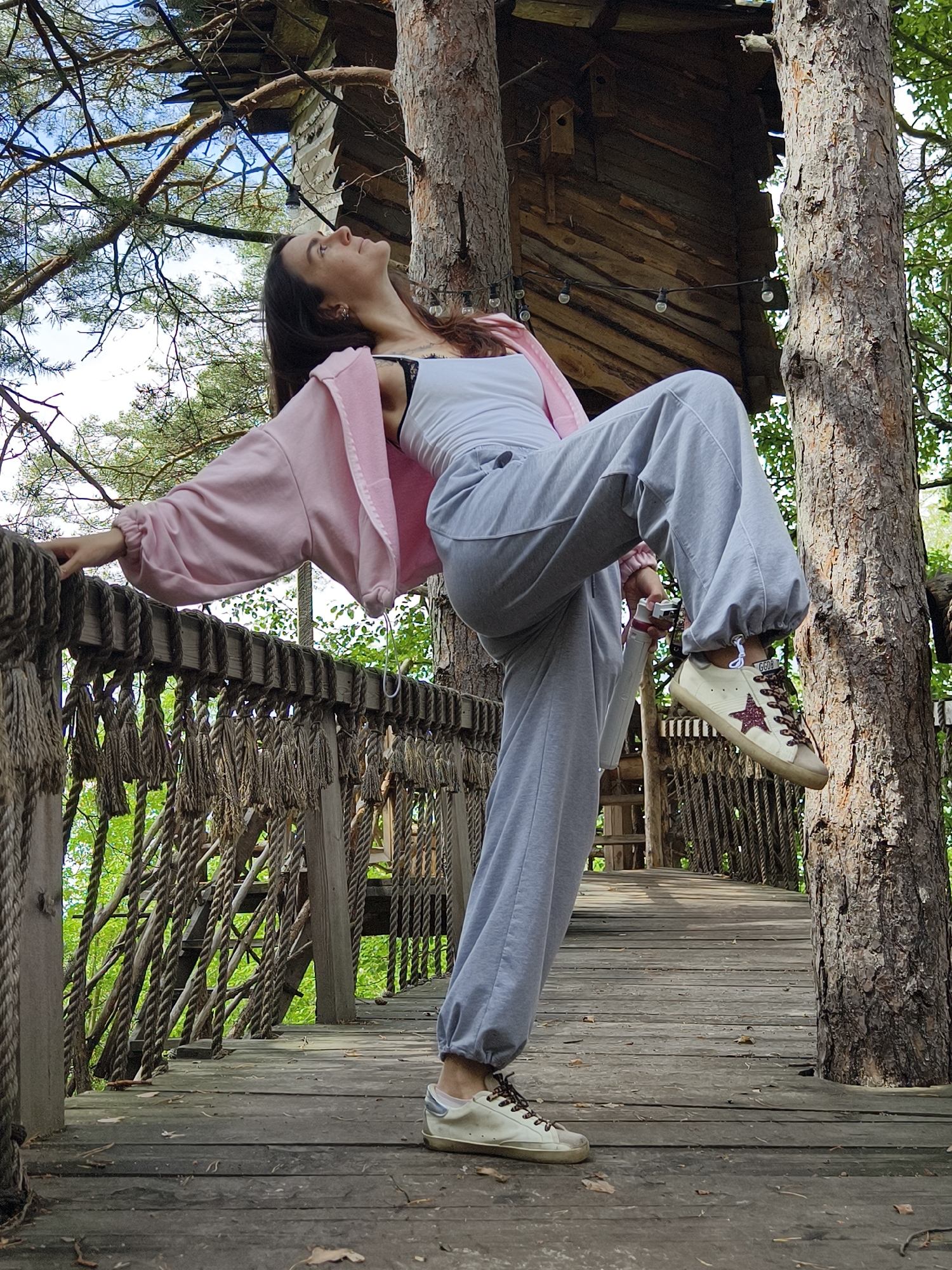 A young woman with long brown hair is posed on a wooden walkway in a treehouse setting. She is wearing a light pink hoodie over a white tank top and grey sweatpants. Her left leg is bent and raised, with her foot resting on the walkway, while her right leg is extended behind her. She is looking upwards, with her head tilted back. Her left hand is resting on a rope railing that borders the walkway. String lights with exposed bulbs are visible, strung between trees and the treehouse structure. The treehouse itself appears to be made of wood and is built amongst tall pine trees. The walkway is also made of wood and has thick rope railings. The scene is outdoors, with lush green foliage visible in the background, suggesting a daytime setting. The woman's pose and upward gaze could suggest contemplation or enjoyment of the surroundings. The location is Onochino, Russia. There is no visible text in the image.