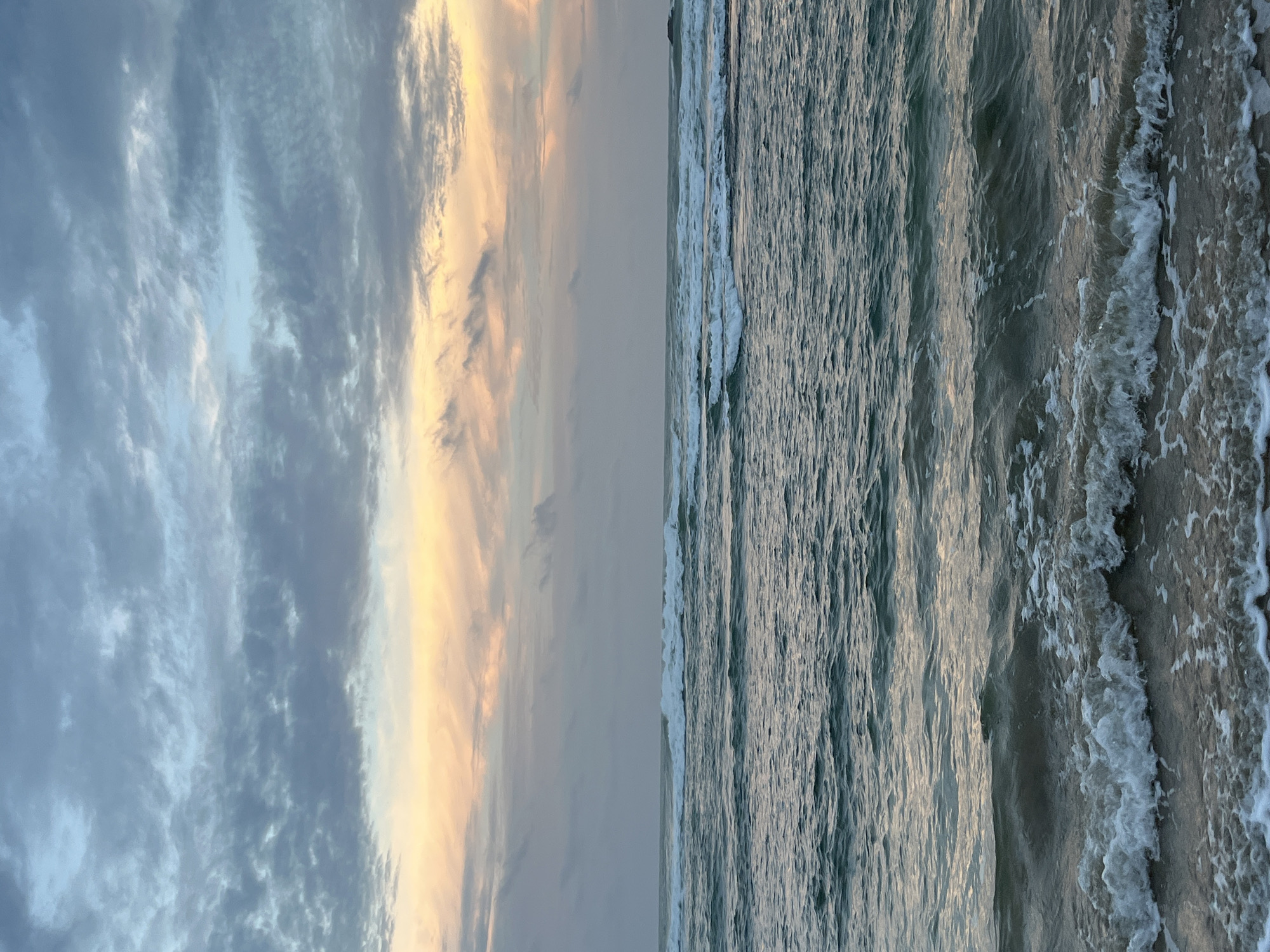 This is a vertical, eye-level shot of a beach at sunset. The image is split down the middle, with the left side depicting the sky and the right side showing the ocean.
The sky is filled with clouds that are illuminated by the setting sun. The clouds on the left are a mix of gray and light blue, with streaks of pale yellow and orange near the horizon. The clouds are wispy and textured, suggesting a gentle breeze.
The ocean on the right side of the image is a deep blue-gray. Gentle waves are rolling in towards the shore, creating white foam and spray as they break on the sand. The water's surface is textured with ripples and patterns. The sand appears wet and dark, reflecting the colors of the sky and water.
The time of day is clearly sunset, as indicated by the warm, golden light hitting the clouds. The weather appears calm and pleasant, with clear skies and gentle waves. The scene evokes a sense of peace and tranquility. No people or objects are visible in the image. The location is identified as Kathaluwa, Sri Lanka. There is no visible text in the media file.