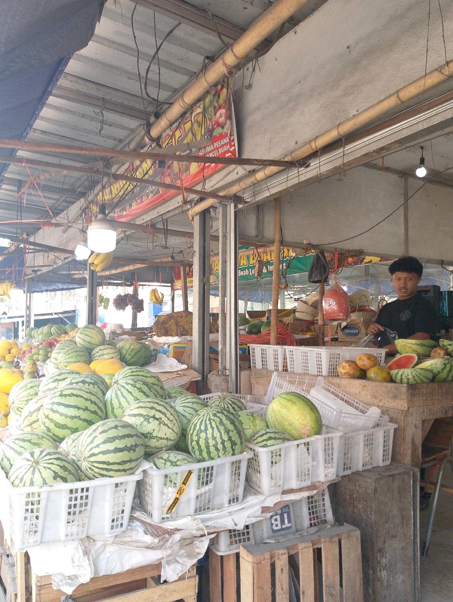 This image captures a vibrant fruit stall in Lembang, Indonesia. In the foreground, an abundant display of watermelons, both whole and cut, fills white plastic crates. To their right, a vendor, a young man with curly hair wearing a black t-shirt, stands behind the counter. He holds a plastic bag and appears to be interacting with a customer or preparing to serve. Behind him, various other fruits like papayas and possibly melons are visible, along with hanging bunches of grapes and bananas. The stall is covered by a corrugated metal roof supported by bamboo poles, with a brightly colored banner advertising fruit hanging above. The scene is bustling with a variety of produce, suggesting a busy market environment.