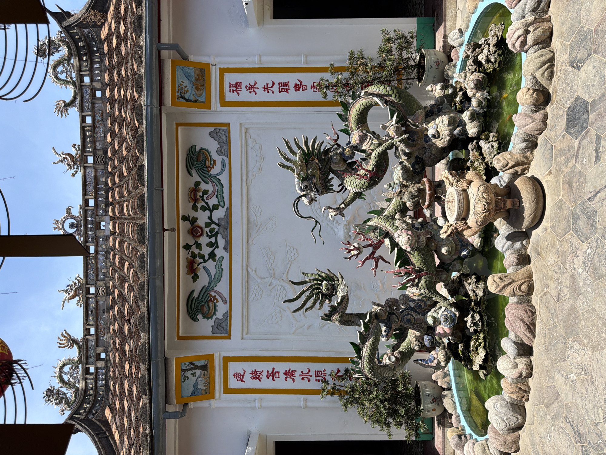 The image shows the exterior of a building with a tiled roof and ornate dragon sculptures. The building has white walls decorated with colorful murals and vertical red inscriptions. In front of the building is a pond with water, rocks, and a large, multi-headed dragon sculpture. There are also potted plants and paved ground. The sky is clear and blue. The location is identified as Hội An Ward, Vietnam.