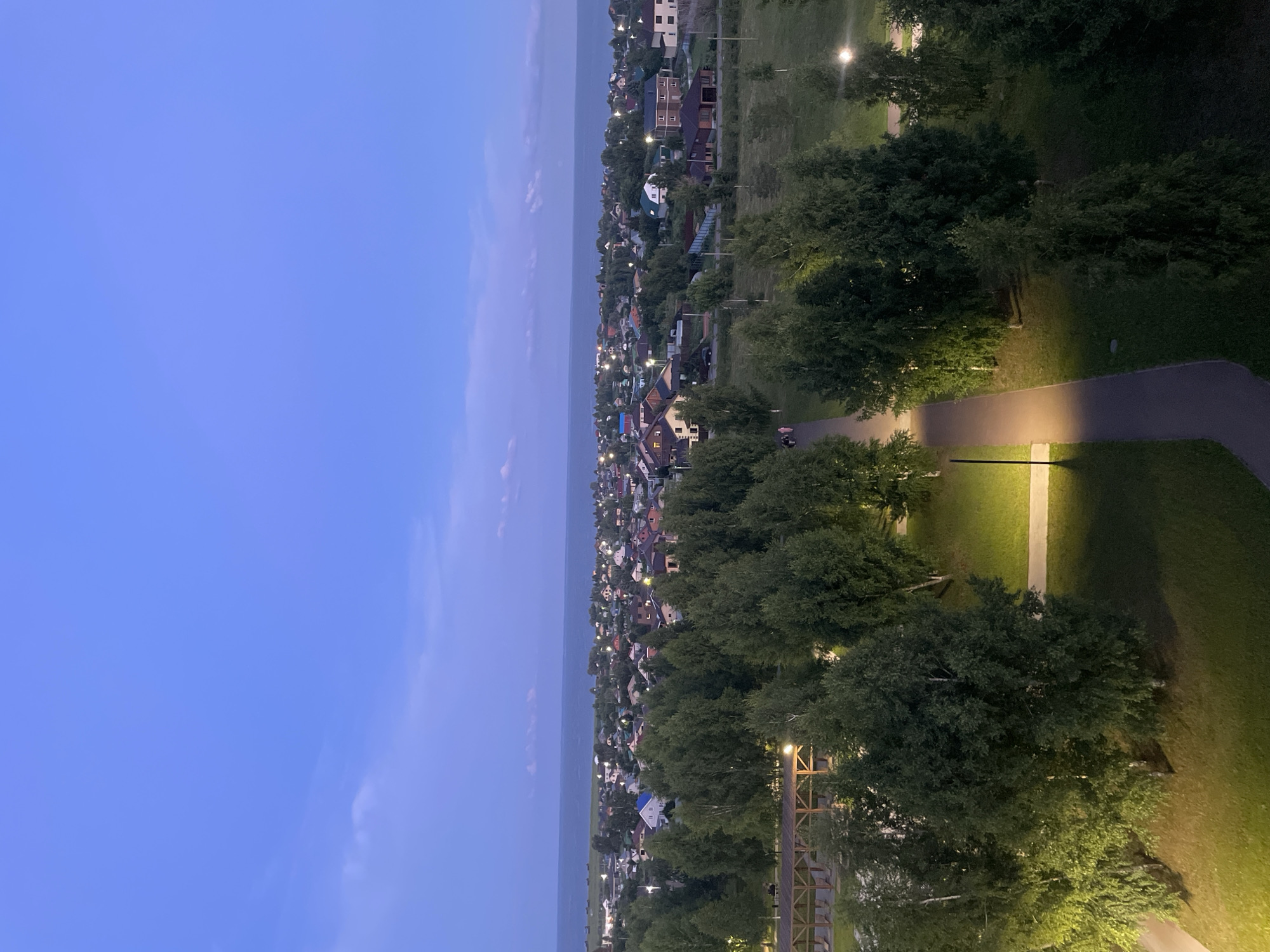 An aerial, wide-angle, nighttime photograph shows a view of a residential area near the water, in Laishevo, Russia. In the foreground, a paved walkway curves through a grassy area with several trees, some illuminated by streetlights. A wooden structure with a roof and seating is visible in the upper left. In the middle ground, a dense cluster of trees separates the park from a residential area with numerous houses of varying sizes and architectural styles, many with illuminated windows. Beyond the houses, a body of water, possibly a lake or sea, stretches to the horizon under a deep blue, twilight sky with a few wispy clouds. The overall scene depicts a tranquil evening in a suburban or semi-rural setting. Two figures are visible walking on the path in the middle ground.
