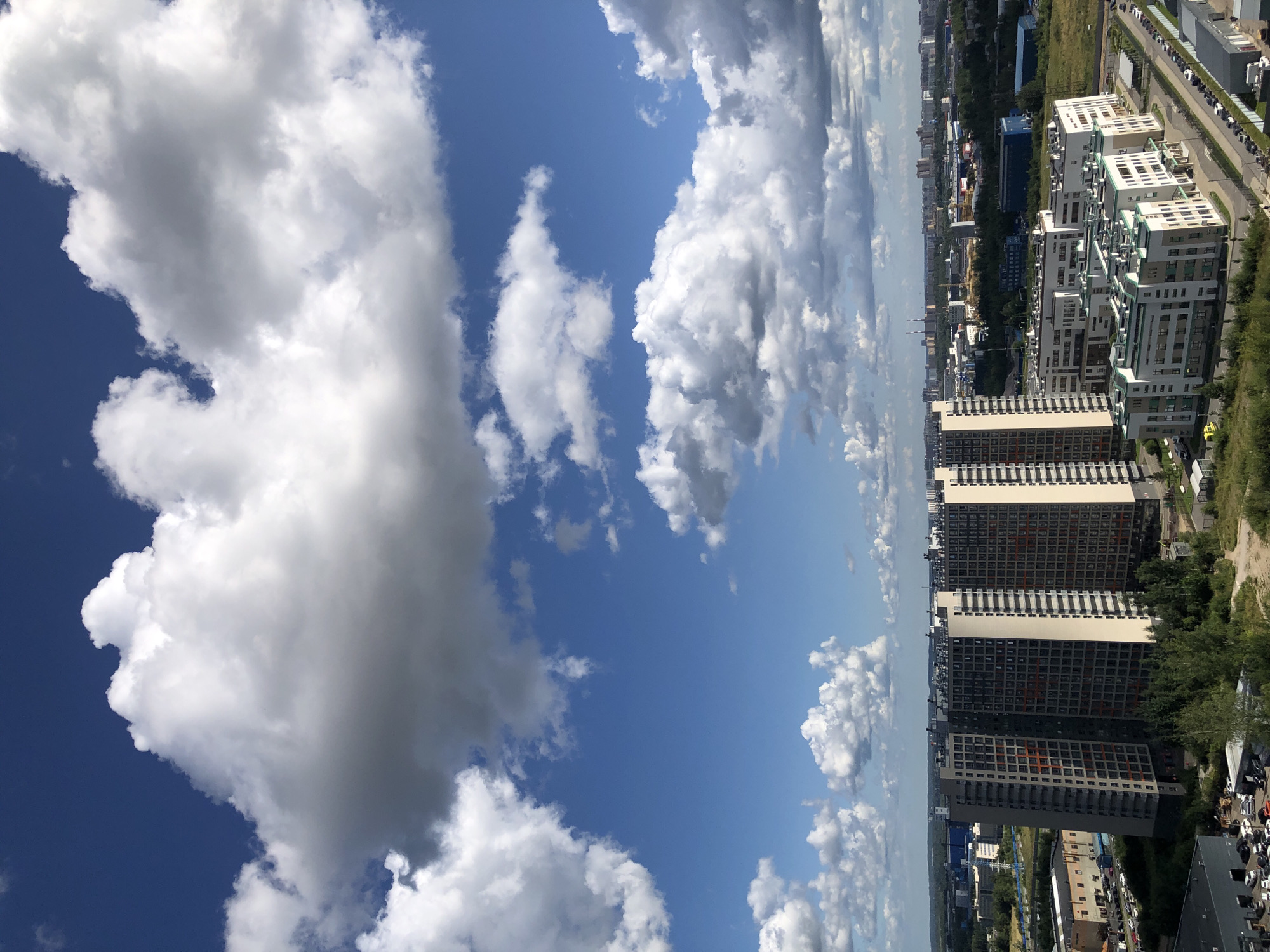 An aerial shot captures a sprawling cityscape under a partly cloudy sky. The foreground is dominated by a cluster of modern, multi-story apartment buildings, characterized by their uniform balconies and contrasting facade colors. To the left, a taller, slender building stands out with its dark gray exterior and orange window accents. Adjacent to it, a series of lower, white buildings with green accents are neatly arranged, facing a paved road lined with parked cars. Beyond the immediate residential area, the cityscape extends with a mix of industrial structures, including warehouses and factories, interspersed with patches of green vegetation. In the distance, a hazy horizon suggests the presence of more urban development and possibly natural landscapes. The sky above is a vibrant blue, punctuated by large, fluffy white and gray clouds that cast shadows over parts of the urban landscape. The scene appears to be an urban or suburban environment with a focus on residential and industrial architecture.