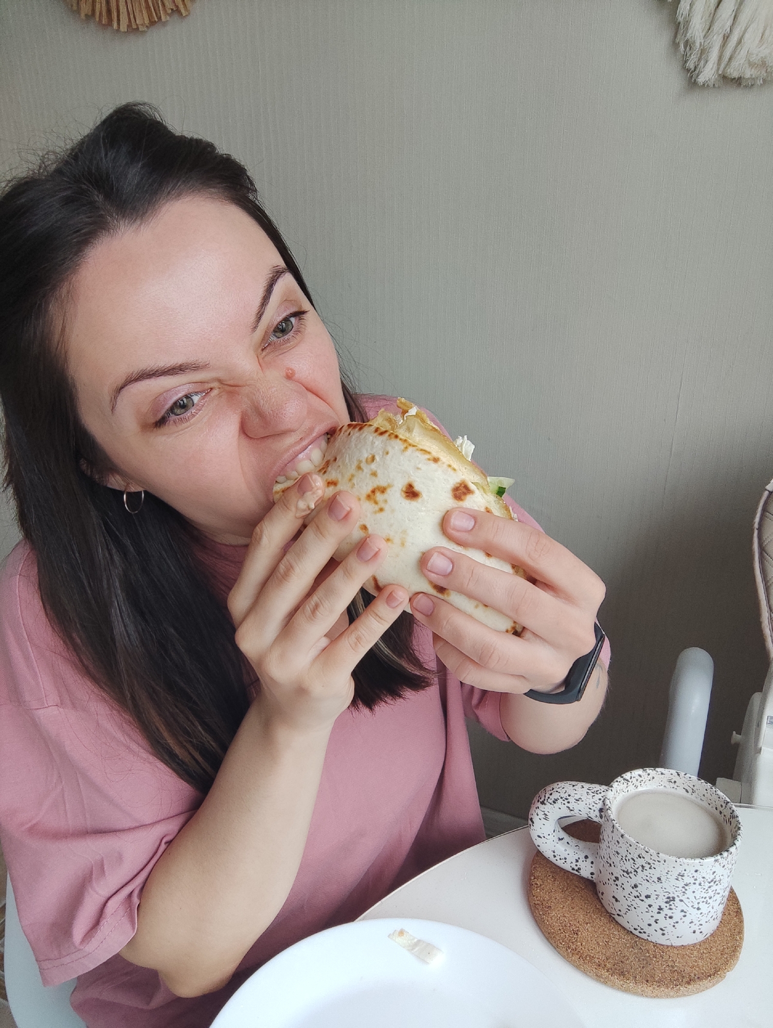 A woman is seated at a table, eating a wrap. She has dark hair pulled back, and is wearing a pink t-shirt. Her expression suggests she is enjoying the wrap. She is holding the wrap with both hands, and has taken a large bite. A mug with a speckled pattern sits on a circular coaster next to a plate. The background is a plain wall with a decorative straw hanging. The location of the scene is Unknown, Unknown.
