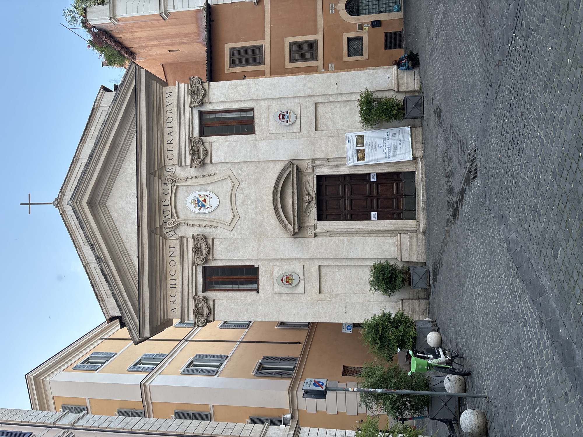 The image depicts a building facade with the inscription "ARCHICONFRATIS CARCERATORVM." The building appears to be a church or religious structure, featuring a prominent entrance door, windows, decorative elements, and a cross on the roof. In front of the building, there's a cobblestone street with trees planted in square containers and a few parked bicycles. Two people are seated on the ground next to the building. The style of the architecture is consistent with that of Rome, Italy.