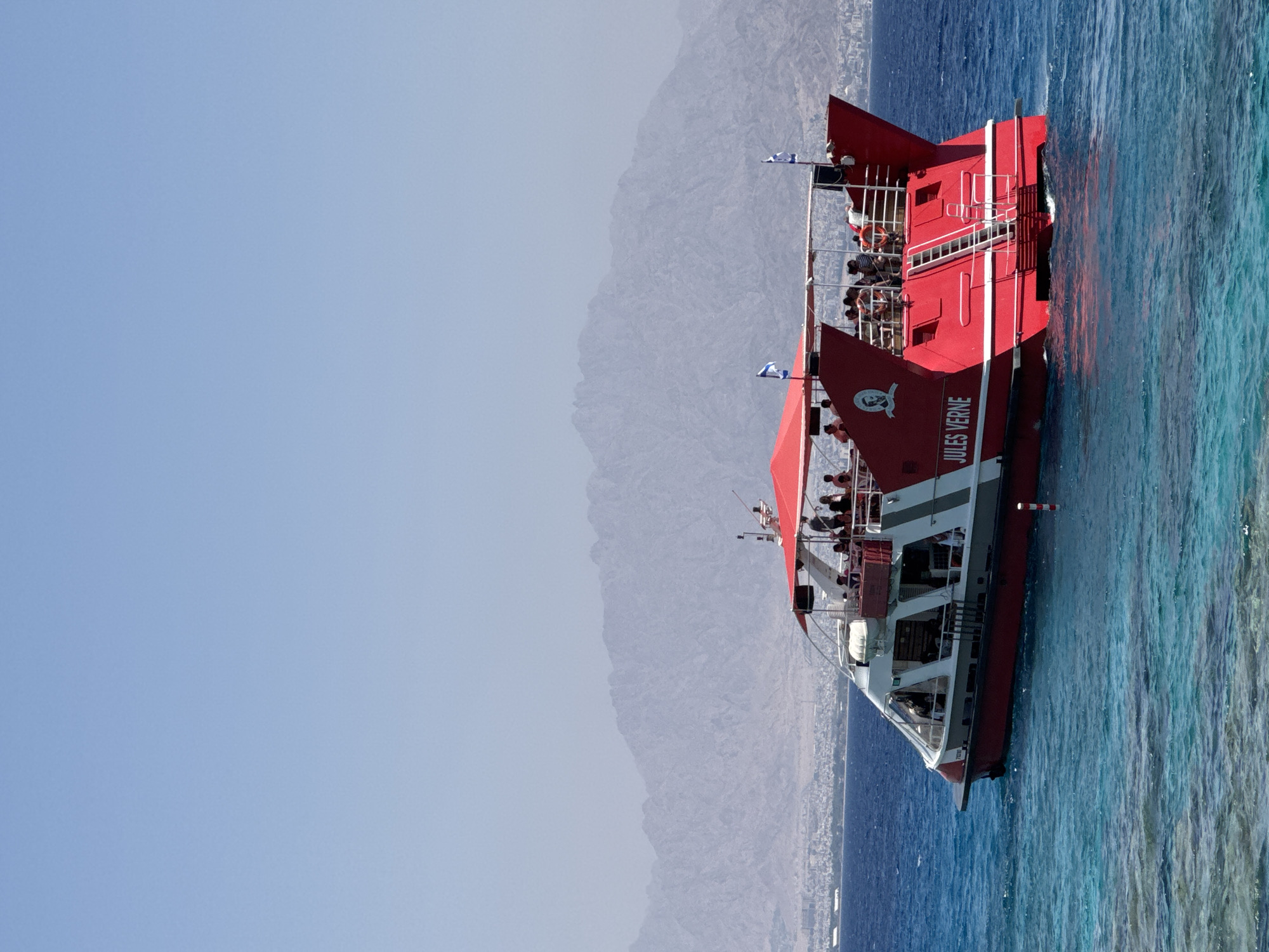 A red and white boat named "JULES VERNE" is seen on the bright blue sea. The boat has multiple decks with people standing and sitting on them. The boat is in motion, as indicated by the wake behind it. In the background, a hazy mountain range is visible, with a city lining the coast below it. This location is likely Eilat, Israel, known for its Red Sea coastline and arid mountain backdrop. The water is clear and shows hints of a coral reef below the surface in the foreground. The sky is a clear, light blue.