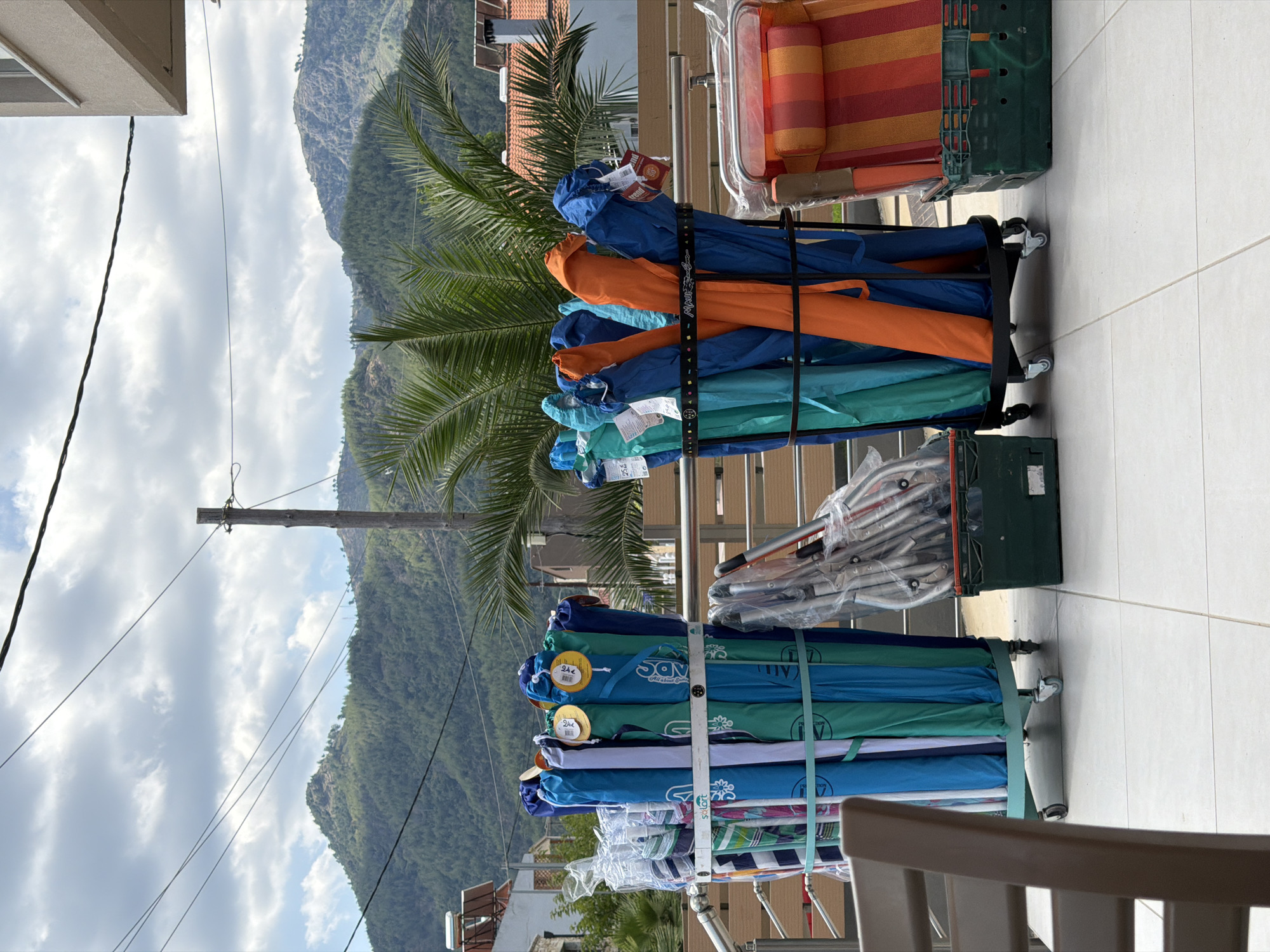 In Skala Potamias, Greece, on a balcony or patio area, a collection of beach umbrellas and chairs are neatly arranged. Two large carts are filled with various colored beach umbrellas, primarily in shades of blue, teal, and orange, some still in their protective plastic packaging. To the right, a striped beach chair with orange and red stripes rests in a green plastic crate. In the background, a lush green mountainous landscape is visible, with scattered buildings and telephone wires overhead. The sky is partly cloudy, with a mix of white clouds and blue sky. The ground is tiled with large, light-colored square tiles. A portion of a wooden railing separates the balcony from the lower area, and a dark brown chair is visible in the lower left corner of the frame. No people are present in the scene.