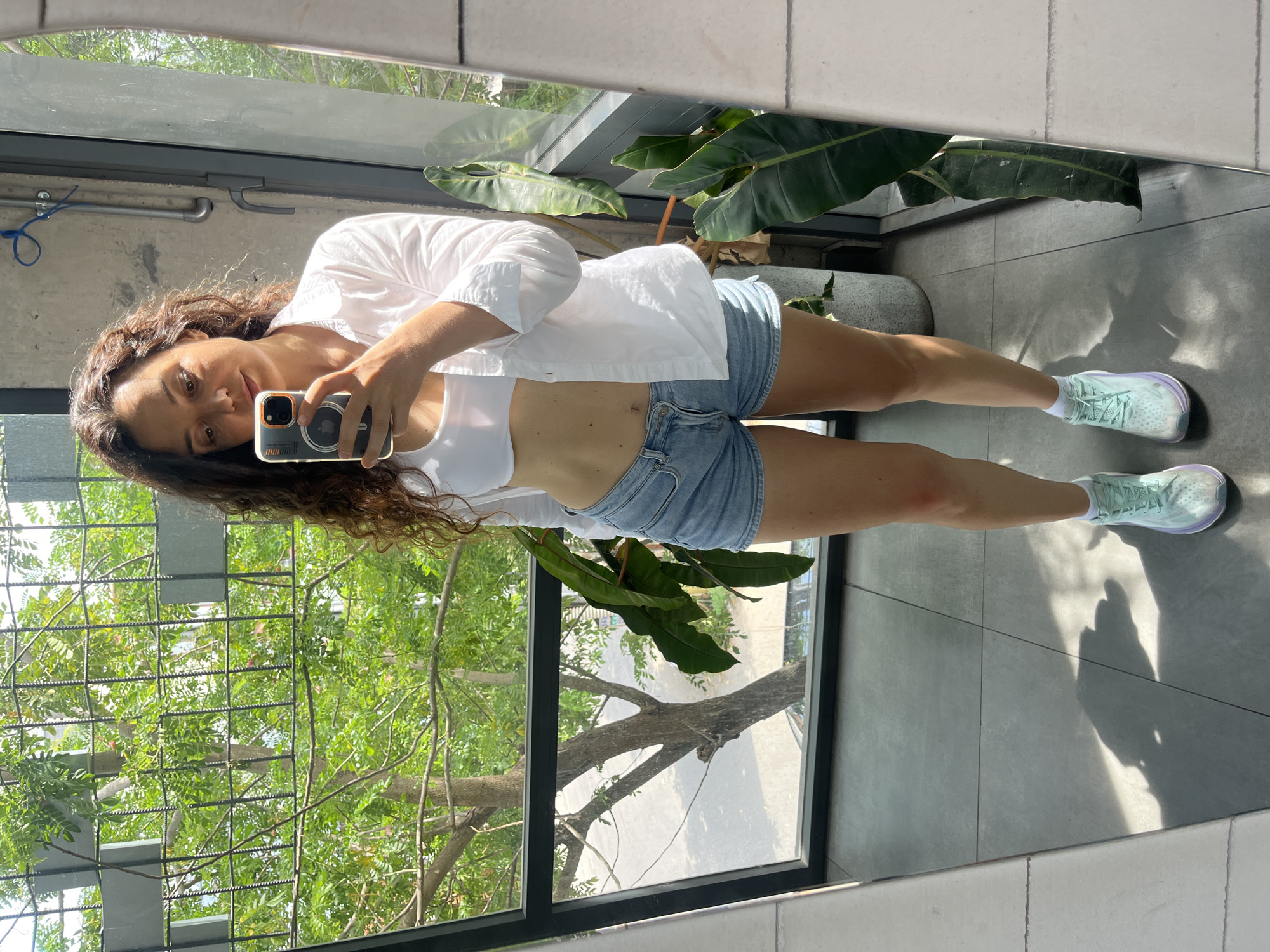 In Phu Thuy Ward, Vietnam, a woman is captured in a full-body mirror selfie taken from a slightly low angle. She stands indoors, facing a large window with a metal grid structure. Outside the window, lush green trees are visible, with a street and parked cars in the background. The woman is wearing a white unbuttoned collared shirt over a white crop top, light blue denim shorts, and mint green sneakers with white laces. Her dark, curly hair falls around her shoulders. She is holding a smartphone in her right hand, angled to take the photo. The environment appears to be a modern interior with concrete walls and grey tiled flooring. Large potted plants with broad green leaves are placed near the window, adding to the natural elements within the scene. Sunlight casts distinct shadows on the floor, suggesting a bright day.