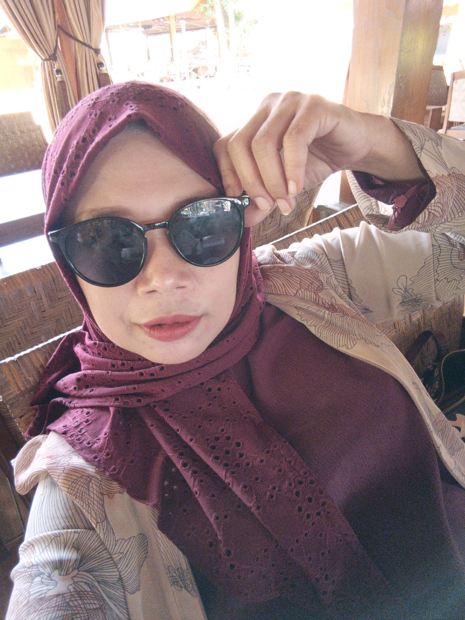 A selfie features a woman wearing a maroon hijab and sunglasses, posing with her hand on her sunglasses. She is seated at a woven rattan table, possibly in an outdoor setting as indicated by the blurred background of trees and what appears to be a building with natural lighting. The woman is wearing a light-colored outer garment with a floral pattern over a maroon inner garment. Her lips are painted with red lipstick. The location is specified as Jonggrangan, Indonesia.
