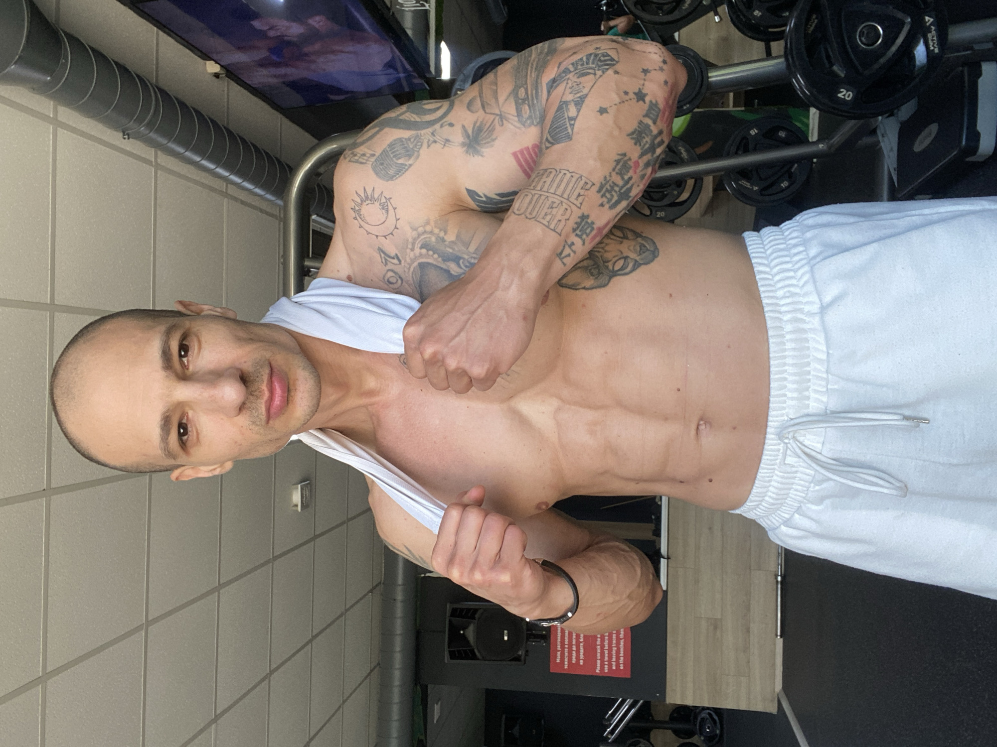 A shirtless man with a shaved head, visible chest muscles, and extensive tattoos on his upper arms stands in a gym setting. He wears white athletic pants and is holding a white tank top. Exercise equipment, including weight plates, is visible behind him. In the background, there is a sign written in a language other than English. The environment appears to be well-lit with tiled walls. The location could potentially be the city of Pleven, Bulgaria, based on the text on the sign.