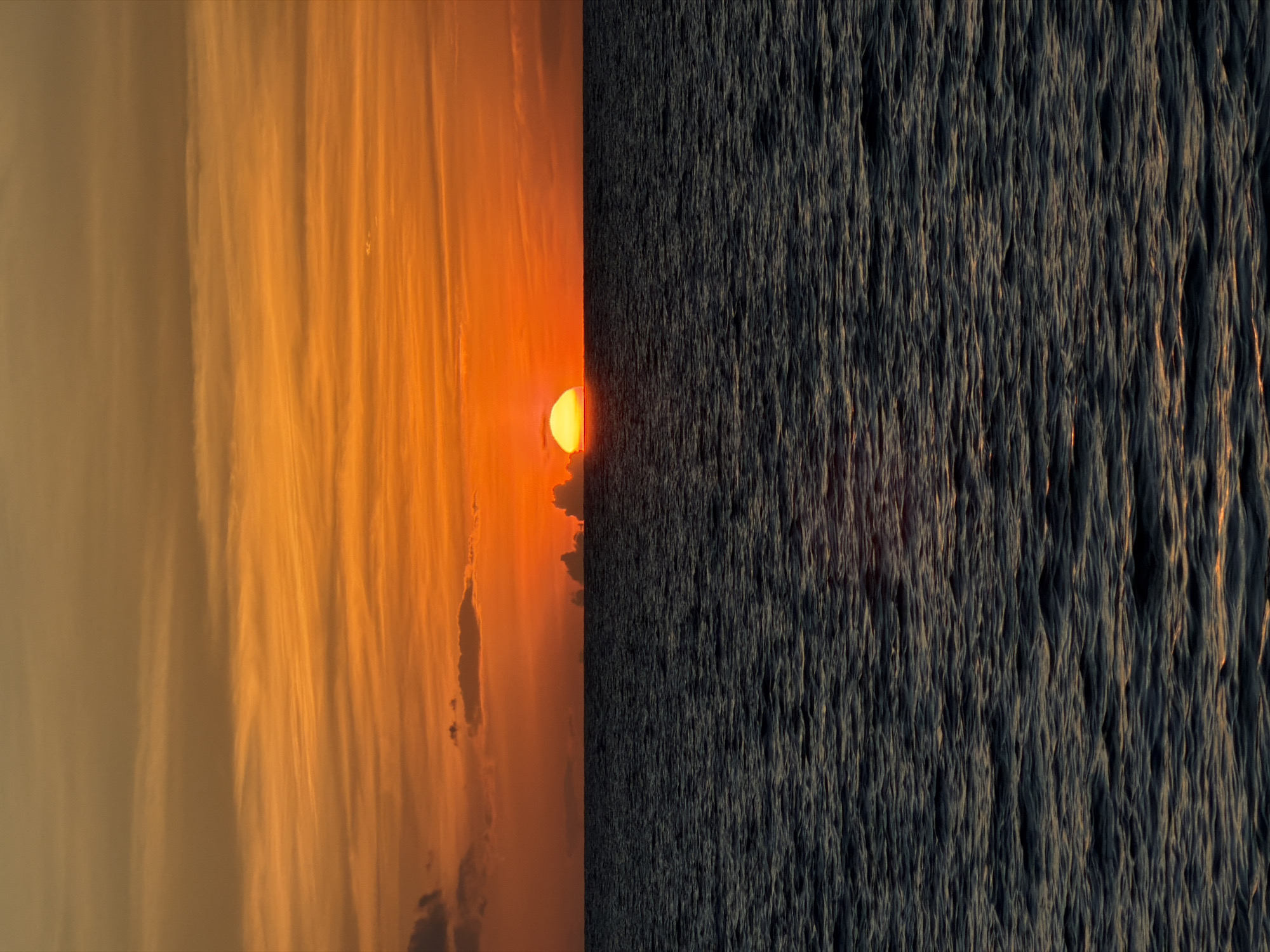 This media file displays a split image, with the left side showing a vibrant sunset over a cloudy sky and the right side depicting choppy, dark water. The sunset is a brilliant orange, casting a warm glow on wispy, elongated clouds that stretch across the frame. The sun itself is a bright, semi-circular disc, partially obscured by the horizon. A few darker clouds are also visible, adding depth and contrast to the sky.
The right half of the image is dominated by a turbulent body of water, likely the sea, given the location context of Baan Plai Laem, Thailand. The water is a deep, dark grey with significant texture, indicating rough waves or currents. Glimmers of light reflect off the crests of these waves, suggesting the fading light of the sunset is still reaching the water's surface, though much less intensely than the sky. The overall impression is one of contrast between the calm beauty of the sky and the dynamic energy of the water.
The time of day is clearly sunset, characterized by the intense orange hues and the low position of the sun. The weather appears to be clear enough for a dramatic sunset, but the choppy water suggests conditions might be windy or have a strong current. The location cues point to a coastal area in Baan Plai Laem, Thailand, likely a beach or overlooking the sea. No people or objects are visible, and there is no visible text. The juxtaposition of the serene sky and the agitated water could evoke a range of emotions, from awe at nature's beauty to a sense of power and perhaps even a hint of foreboding from the dark, churning sea.