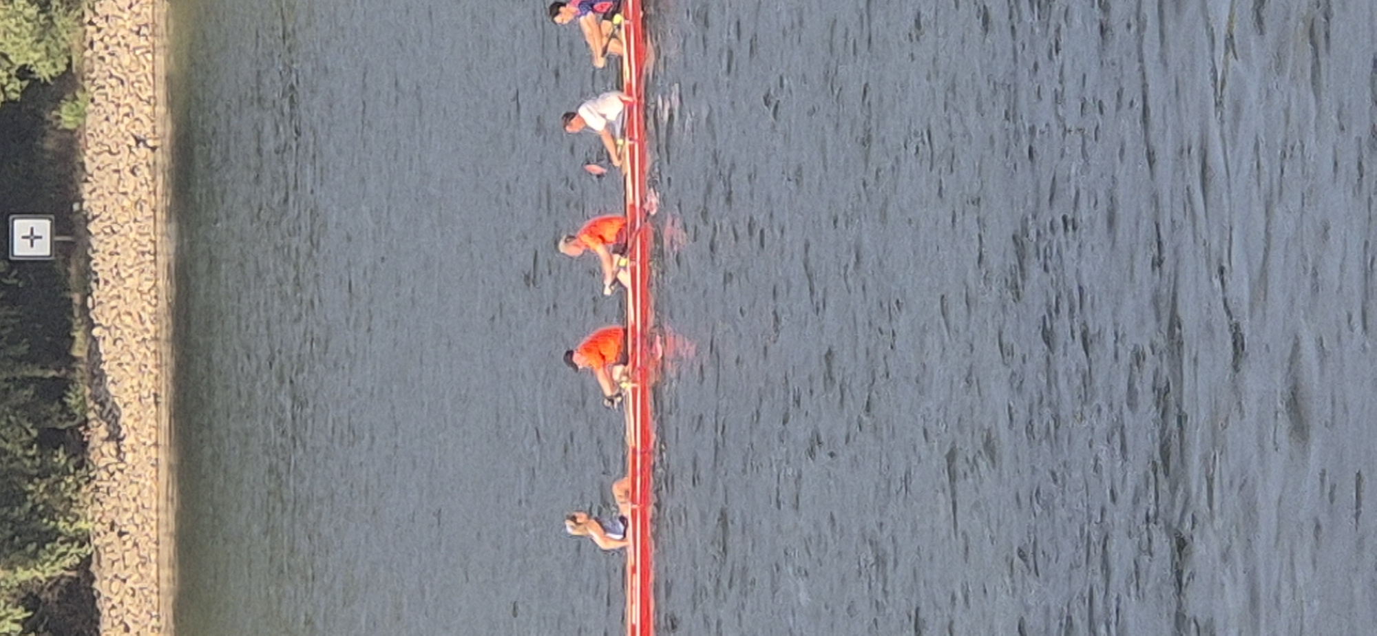This is a high-angle, full-shot photograph of a dragon boat racing on the Rhine River in Mainz, Germany. The boat is red with a white stripe and is occupied by five individuals. The individuals are engaged in rowing, with four men and one woman. The men are wearing orange shirts, while the woman is wearing a blue and white top. The river is calm, with ripples on the surface. In the background, a rocky shore is visible, lined with trees. A white sign with a plus symbol is present on the shore, likely indicating a marker. The lighting suggests it is daytime, possibly late afternoon, with the sun casting reflections on the water. The overall scene depicts a recreational or competitive rowing activity.