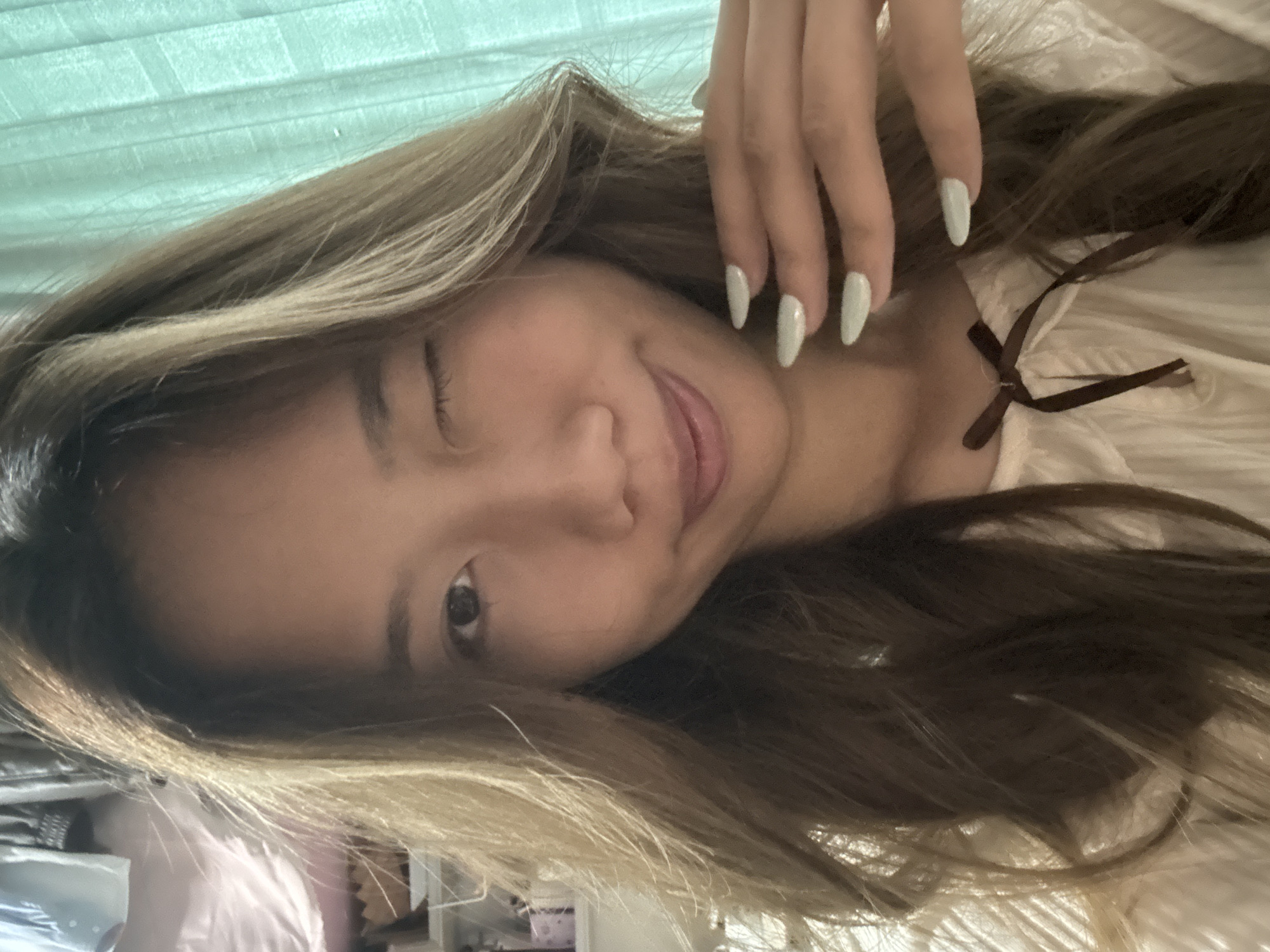 A young woman with long brown hair and blonde highlights poses for a selfie. She is slightly angled with her right eye closed. Her left hand with light green nails is raised near her face. She is wearing a light-colored top with a brown ribbon at the neckline. A green background is partially visible behind her. A cluttered area with various objects is visible in the lower left corner. The location in the city of Nan'an District, China is not discernible from the image.