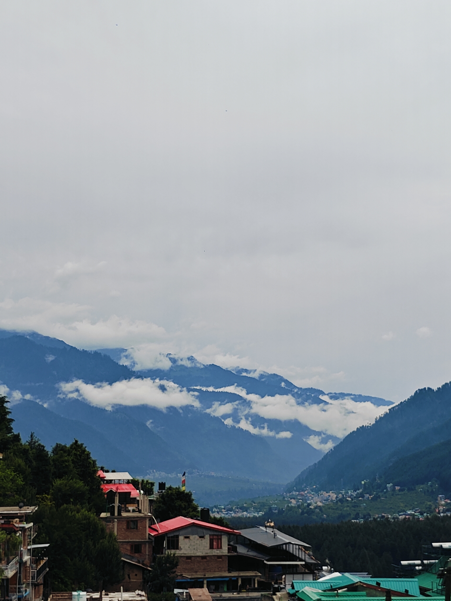 This media file captures a scenic view of the Himalayan mountains in Vashisht, India, under a cloudy sky. The image shows layers of lush green mountains interspersed with white clouds, creating a dramatic and atmospheric landscape. In the foreground, there are buildings with red and green roofs, indicating a settlement nestled on the hillside. Some of these buildings appear to be residential or commercial structures, with visible windows and balconies. A flag is also visible on one of the buildings. The overall scene suggests a tranquil setting in a mountainous region, possibly during the daytime given the ambient light. The weather appears to be overcast, with the clouds obscuring parts of the mountain range and creating a soft, diffused light. The location cues, such as the style of the buildings and the mountainous terrain, strongly suggest a place like Vashisht, known for its scenic beauty and proximity to the Himalayas. There are no visible people in the image, and the focus is on the natural landscape and the settlement. The image evokes a sense of peace and awe, characteristic of mountain scenery.