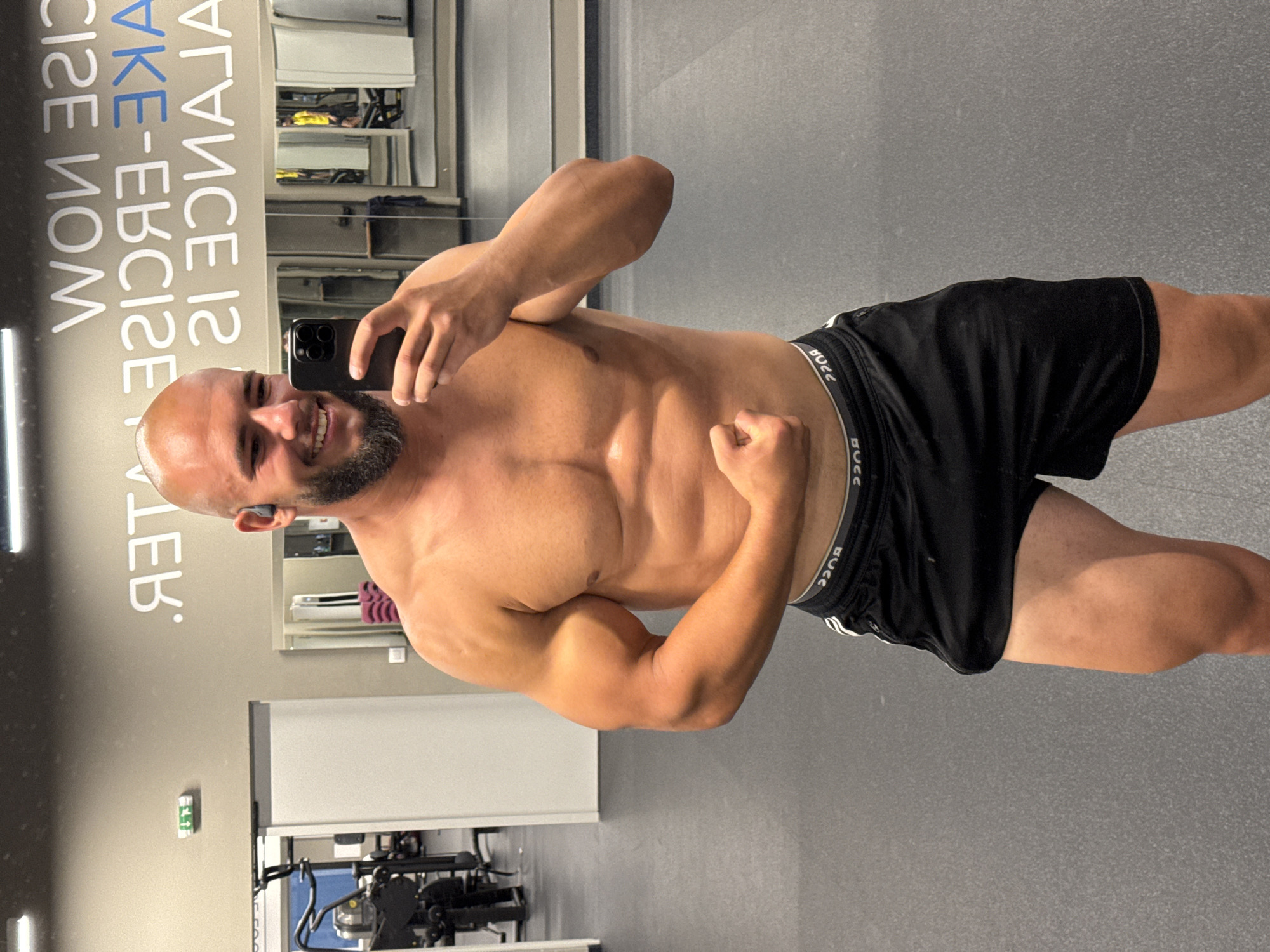A muscular, bald man with a beard is taking a selfie in a gym. He is wearing black shorts. The gym appears to be in Sofia, Bulgaria, as indicated by the text on the wall behind him. The text, which is backwards, reads "WON 3212 3HIA-380132 21 30ИAJAU" and translates to "NOW EXERCISE-TAKE BALANCE IS 21". The man is flexing his bicep. In the background, there are gym equipment, mirrors, and other people exercising. The floor is dark gray.