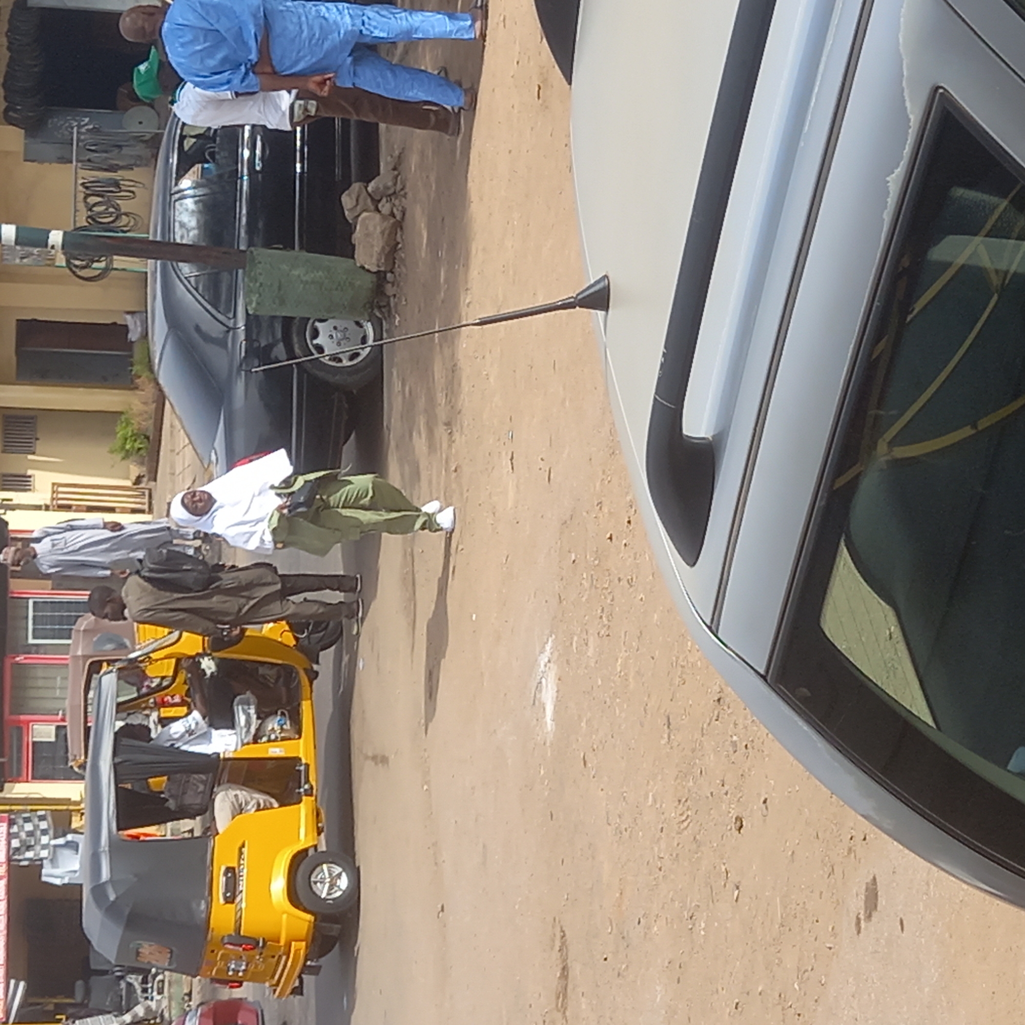 This daytime image, captured in Sardauna, Nigeria, portrays a lively street scene with several individuals and vehicles on a light brown, sandy surface.
In the upper left portion of the frame, two men are standing near a black sedan. One man wears a light blue traditional attire, while the other is dressed in a white shirt and brown trousers. A third person in a green shirt is partially visible behind them. A green post or barrier stands prominently in front of the black car, which has a silver wheel partially visible.
The right side of the image is largely occupied by a gray or silver sedan, showing its roof and side windows. A slender, black antenna-like object protrudes from its roof. Inside the car, seen through the window, are yellow strap-like patterns, possibly from seat covers or items within.
Towards the lower left, a bright yellow tricycle, known locally as a "keke napep," is a central feature. It has a black fabric roof, and several passengers are visible inside. Standing near the tricycle is a man in a dark jacket, carrying a backpack, and another man in a light-colored traditional outfit. In the mid-ground, a person wearing a white top, light green trousers, and white shoes stands, possibly a woman wearing a head covering.
The scene suggests routine daily activities in a populated area, with people gathered and vehicles present. The clear lighting indicates it is daytime. No discernible text is visible on any signs or vehicles.