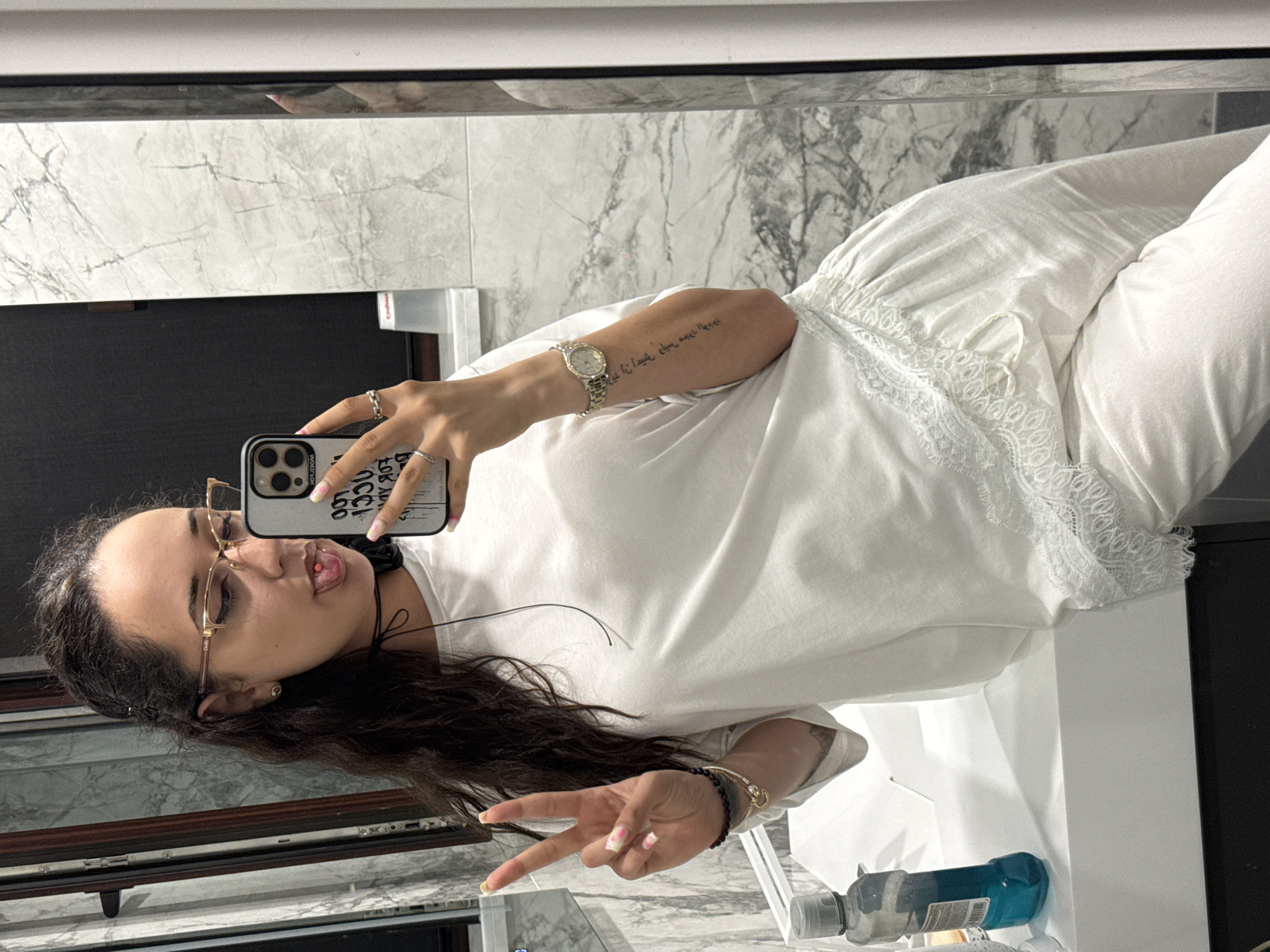 A young woman with dark hair and glasses is taking a mirror selfie in a bathroom. She is wearing a white t-shirt and white pants, both with lace trim. She is sticking her tongue out and making a peace sign with her left hand. A cell phone is visible in her right hand, and she is wearing a watch and bracelets. The bathroom features marble-tiled walls and a white sink.