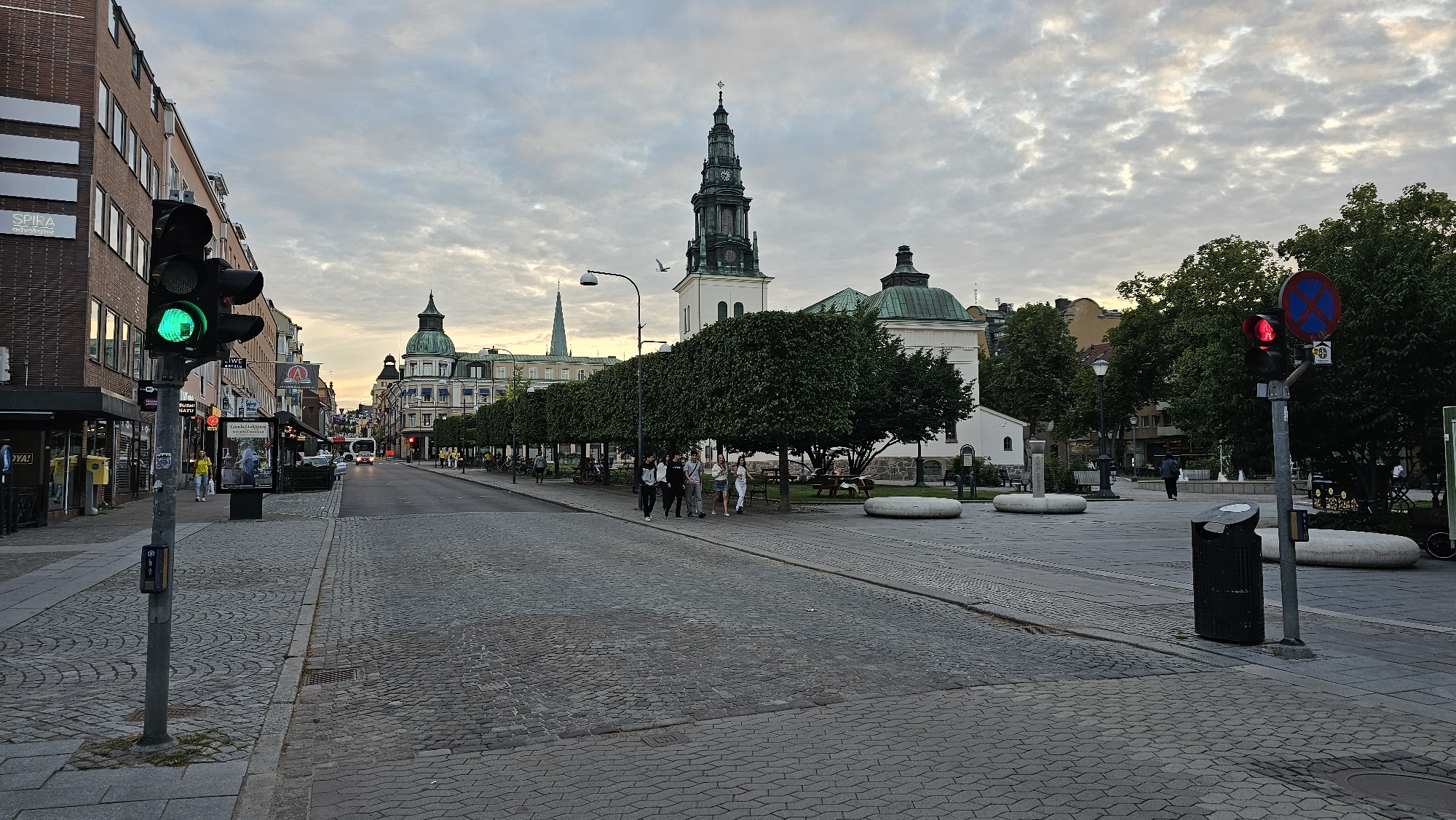 The image depicts a street scene in Linköping, Sweden. A traffic light, currently showing green, is positioned on the left side of the frame, alongside a pedestrian crossing signal. A brick building with multiple windows lines the left side of the street. Further down the street, a church with a tall green steeple is visible in the background, flanked by other buildings with green domed roofs. Several trees with dense green foliage are planted along the sidewalk. A group of five people, four women and one man, are walking on the right side of the street. Another person is walking away from the camera in the distance on the right. The street is paved with cobblestones, and a trash can is visible on the right. The sky is overcast with scattered clouds.