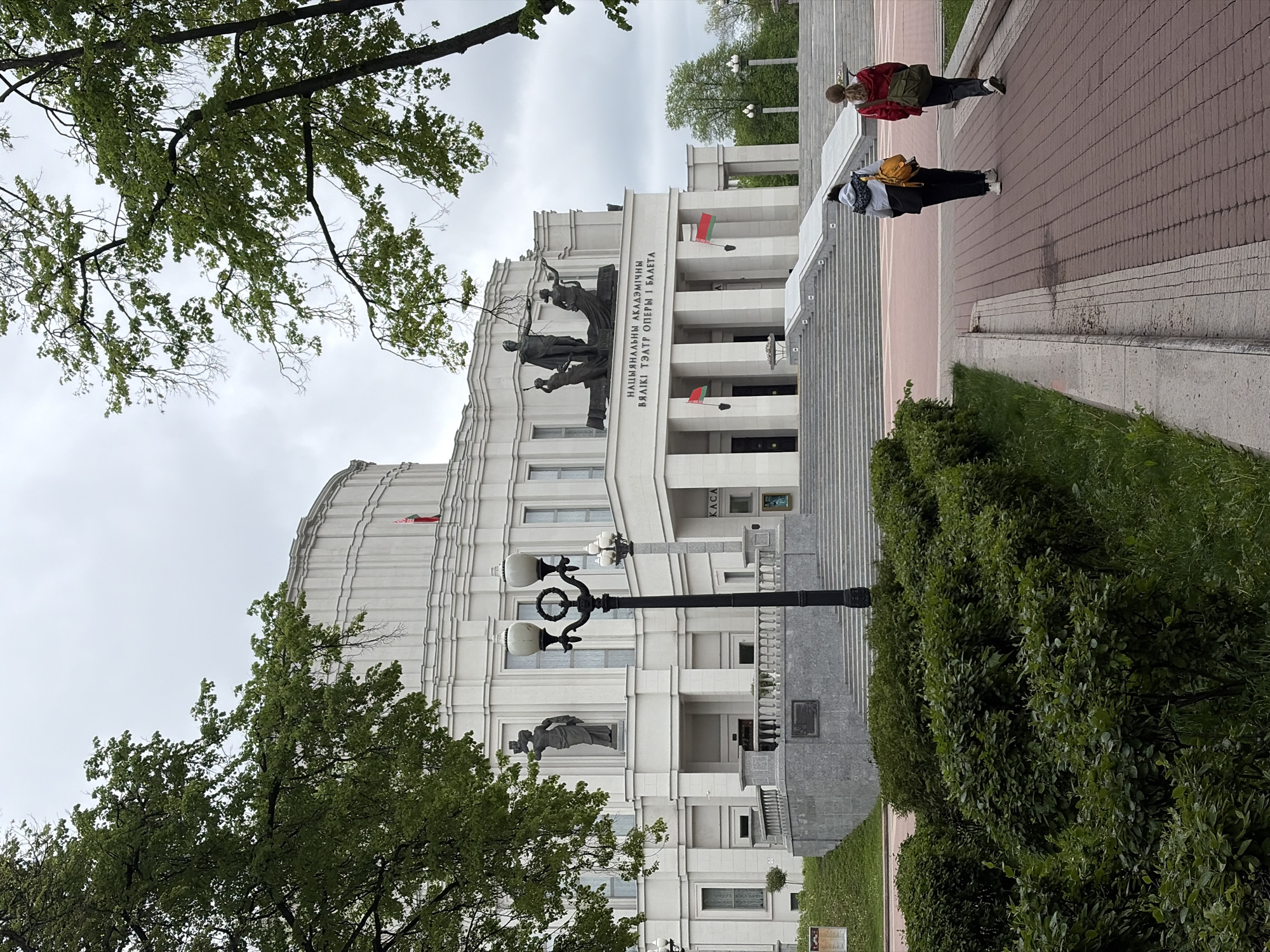 The image shows a building with white façade, ornate details, and flags. On the top, there are sculptures of people on it. The building is the National Academic Grand Opera and Ballet Theatre in Minsk, Belarus. There are people walking on the pedestrian path. To the left, there are trees with green leaves. To the right, there are green bushes. The sky is overcast.