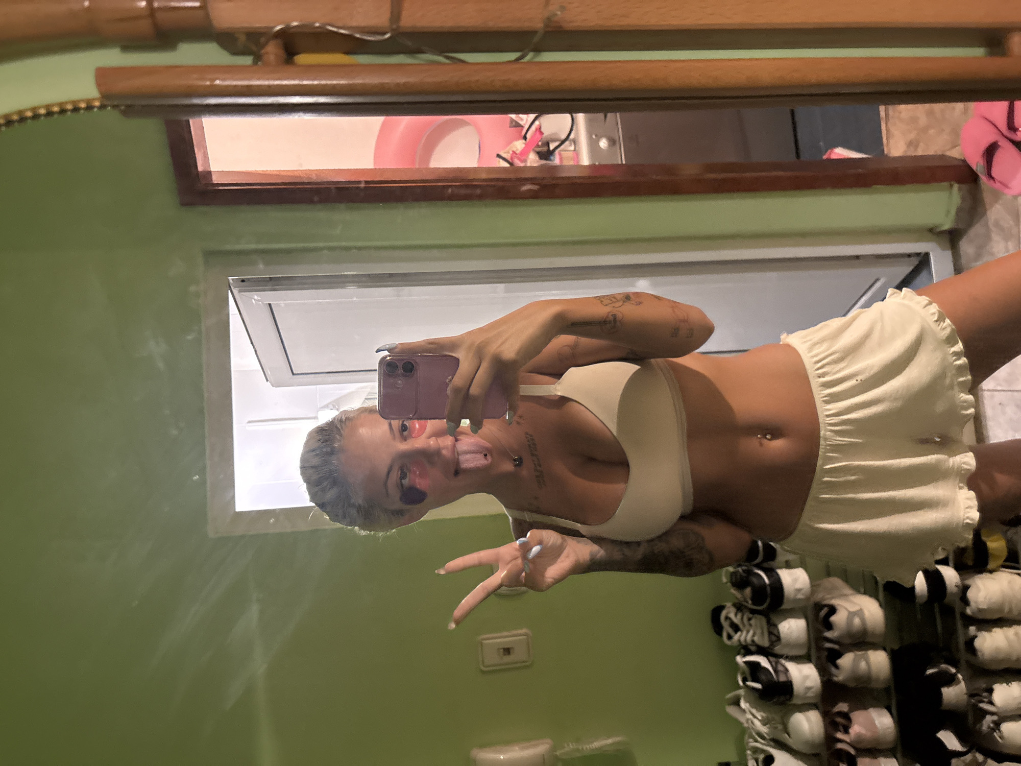 In what appears to be a bathroom or dressing area, a young woman with blonde hair pulled back is taking a mirror selfie. She is wearing a beige bralette and matching ruffled shorts. She has heart-shaped eye patches on and is sticking her tongue out, holding up two fingers in a peace sign. The background shows a green wall, a doorway leading to another room, and a shelf filled with shoes. A mirror reflects the scene.