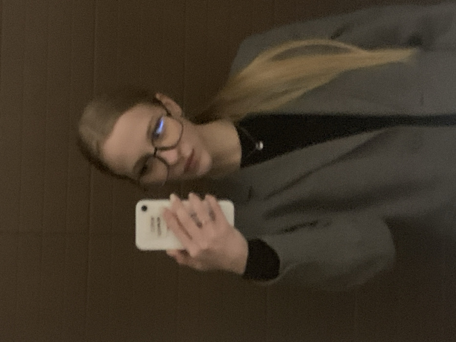 A person with blonde hair and glasses takes a selfie in a mirror. They're wearing a grey blazer and a black top. The phone is white, held in the person's left hand. The background behind the mirror appears to be a plain, brown wall or surface. The overall image is slightly blurred and tilted.