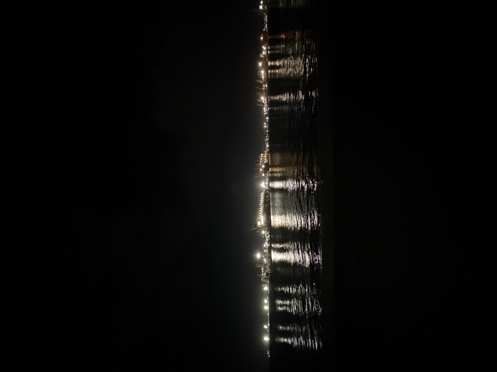 The image shows a night view of a pier or waterfront area in Burgas, Bulgaria. It captures the lights reflecting on the water's surface. There are no people visible. The focus is on the illuminated structure and its reflection, which create a blurry, indistinct pattern on the water. The rest of the image is predominantly dark due to the nighttime setting.