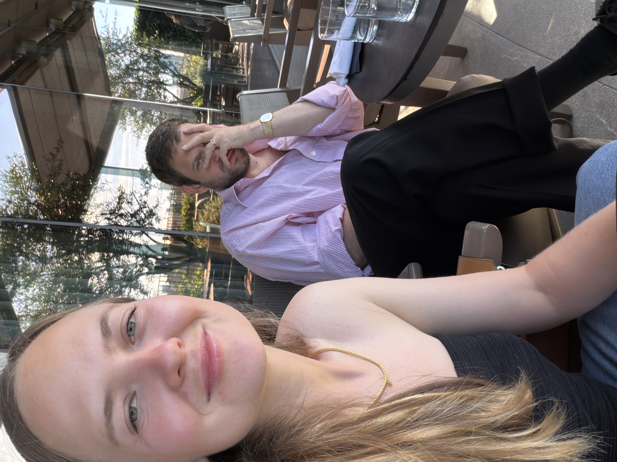 A young woman with blonde hair and green eyes is in the foreground of the image, smiling at the camera. She is wearing a black strapless top and a gold necklace. To her right, a man with brown hair and a beard is seated. He is wearing a pink and white striped button-down shirt and black pants. His left hand is resting on his forehead, and he is wearing a watch on his left wrist. They are seated at a table outside a restaurant with large glass windows reflecting trees and the sky. The scene appears to be a sunny day. The location is identified as Milan, Italy.