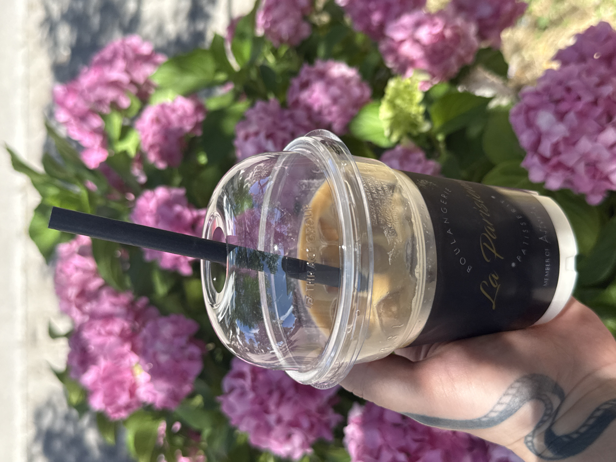 A close-up shot shows a hand with a snake tattoo holding a clear plastic cup of iced coffee. The cup has a black lid and a black straw, with the words "BOULANGERIE La Parisienne PATISSERIE" and "MEMBER OF ASTORIA" printed on a dark label. The background is a blur of vibrant pink hydrangeas, suggesting a setting in a garden or park. The lighting indicates it is daytime, likely outdoors. The image does not provide specific information about the city of Bar, Montenegro, beyond what might be inferred from the presence of hydrangeas, which are common in the region.