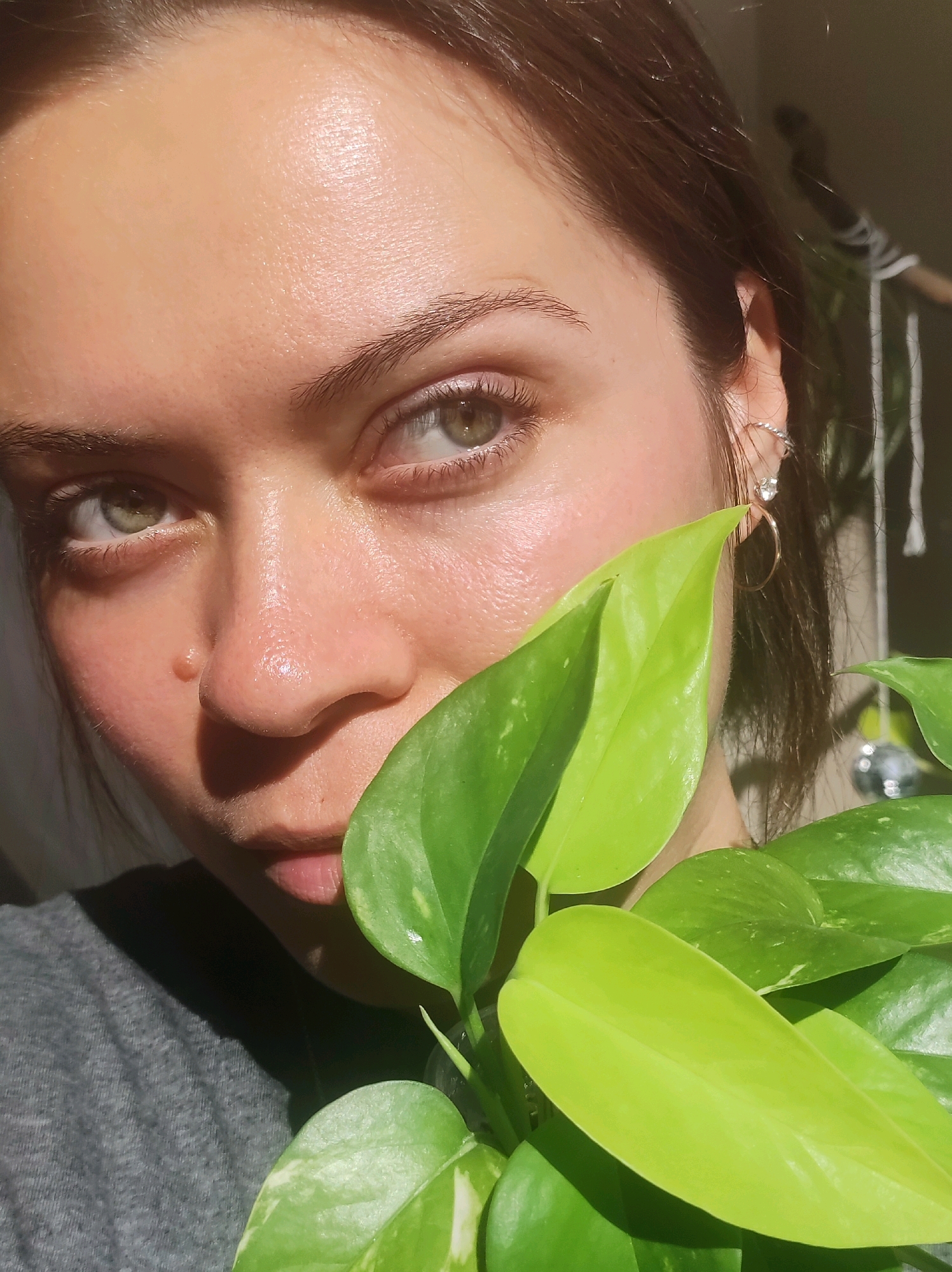 The image is a close-up of a young woman's face, with a plant obscuring part of her nose and mouth. She has light skin, green eyes, and brown hair. She is wearing a gray t-shirt and hoop earrings. The background is blurred but appears to be indoors, with what looks like macrame hanging in the distance. There is no indication of the location being Chaikovsky, Russia.