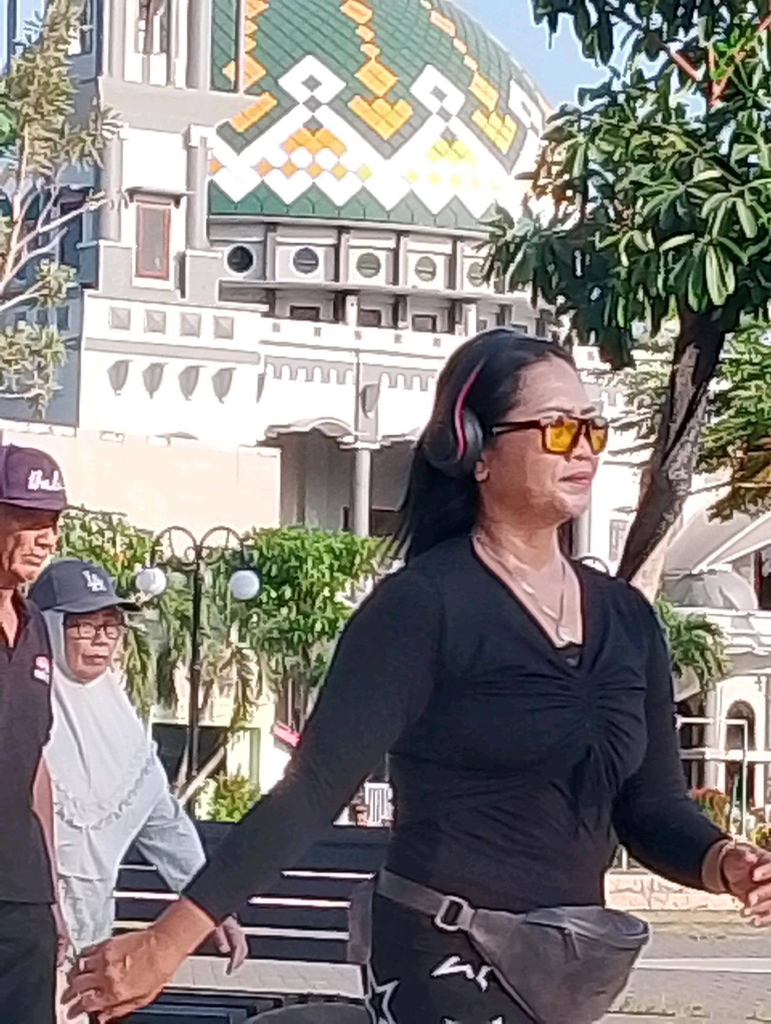 The image shows a group of people near a building with a green and yellow domed roof in Probolinggo, Indonesia. A woman in the foreground, wearing a black long-sleeved shirt, yellow sunglasses, and headphones, is walking to the right. She has a gray fanny pack around her waist. To her left, two other individuals are visible. One person on the far left is wearing a dark shirt and a blue baseball cap. Another person, a woman wearing a white hijab and a light-colored shirt, is in the middle, also wearing a dark baseball cap. The background features a large, ornate building with architectural details and trees. The lighting suggests it is daytime.