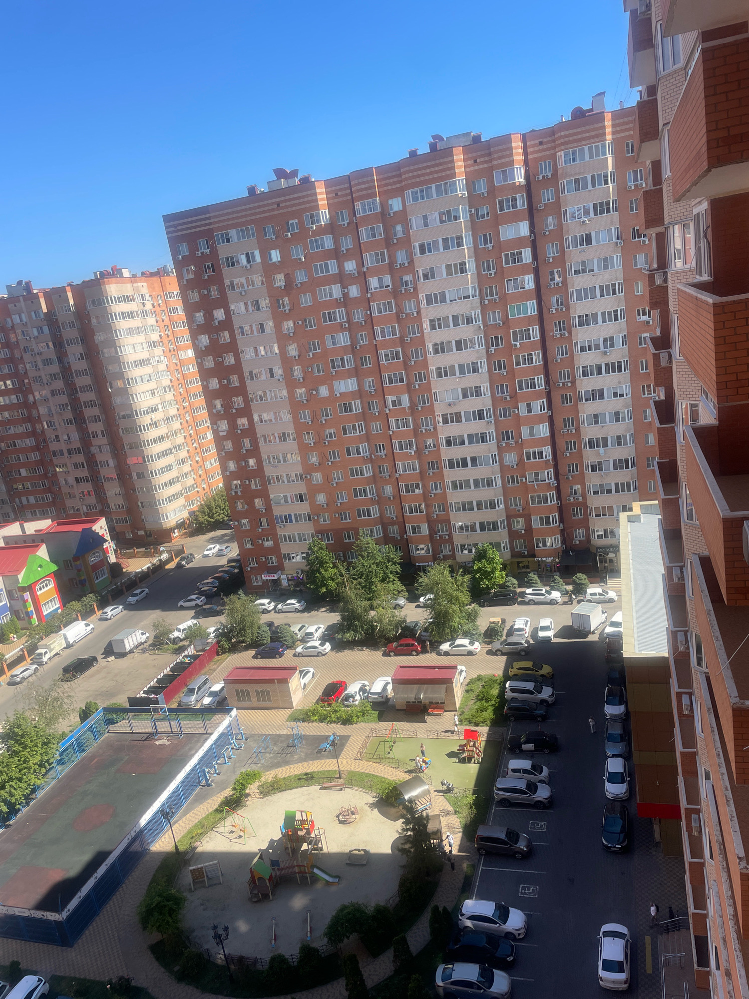 The image captures a high-angle view of a residential complex in Krasnodar, Russia, on a sunny day with a clear blue sky. The scene is dominated by tall, multi-story apartment buildings constructed with red brick and light-colored facades. These buildings are densely packed, suggesting an urban or suburban residential area.
In the foreground and mid-ground, a children's playground is visible, featuring swings, slides, and climbing structures. Adjacent to the playground is a fenced sports court, possibly for basketball or volleyball, with a worn playing surface. Several cars are parked in designated parking areas and along the streets surrounding the buildings. The vehicles are a mix of sedans, SUVs, and vans, in various colors including white, silver, black, and red.
The environment includes paved roads, sidewalks, and green spaces with trees and bushes. The architecture of the buildings suggests modern construction, with numerous windows and balconies. One smaller building with a colorful facade and a red roof appears to be a community facility or a kindergarten. The overall impression is one of a developed residential area with amenities for its inhabitants. There are a few people visible in the image, some walking or standing near the playground and parking areas.