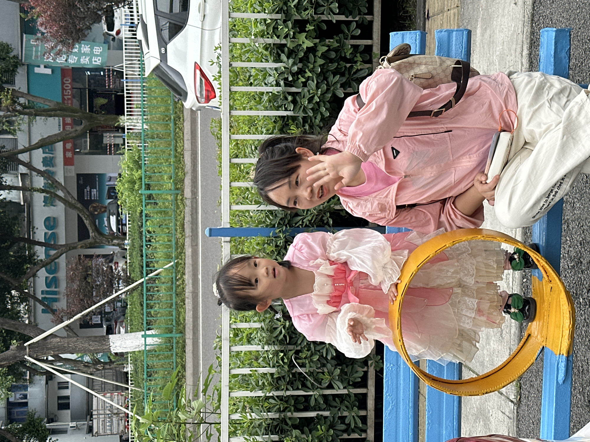 Two young girls are sitting on a blue bench outdoors. One girl is wearing a pink jacket, a light pink shirt underneath, and white pants. She has a beige and brown patterned handbag next to her. She is holding up her hand in a waving gesture. The other girl is wearing a pink dress with white frills. She is positioned behind a yellow hoop. In the background, there is a green hedge and a white fence. Further in the background, there is a street with buildings. A sign on one of the buildings shows the characters “Hisense” (海信), along with the phrase “家电 日接 1500” (Home appliances daily receive 1500). In the background there is a white car. The scene is likely in Nan'an District, China, given the Chinese characters on the sign.