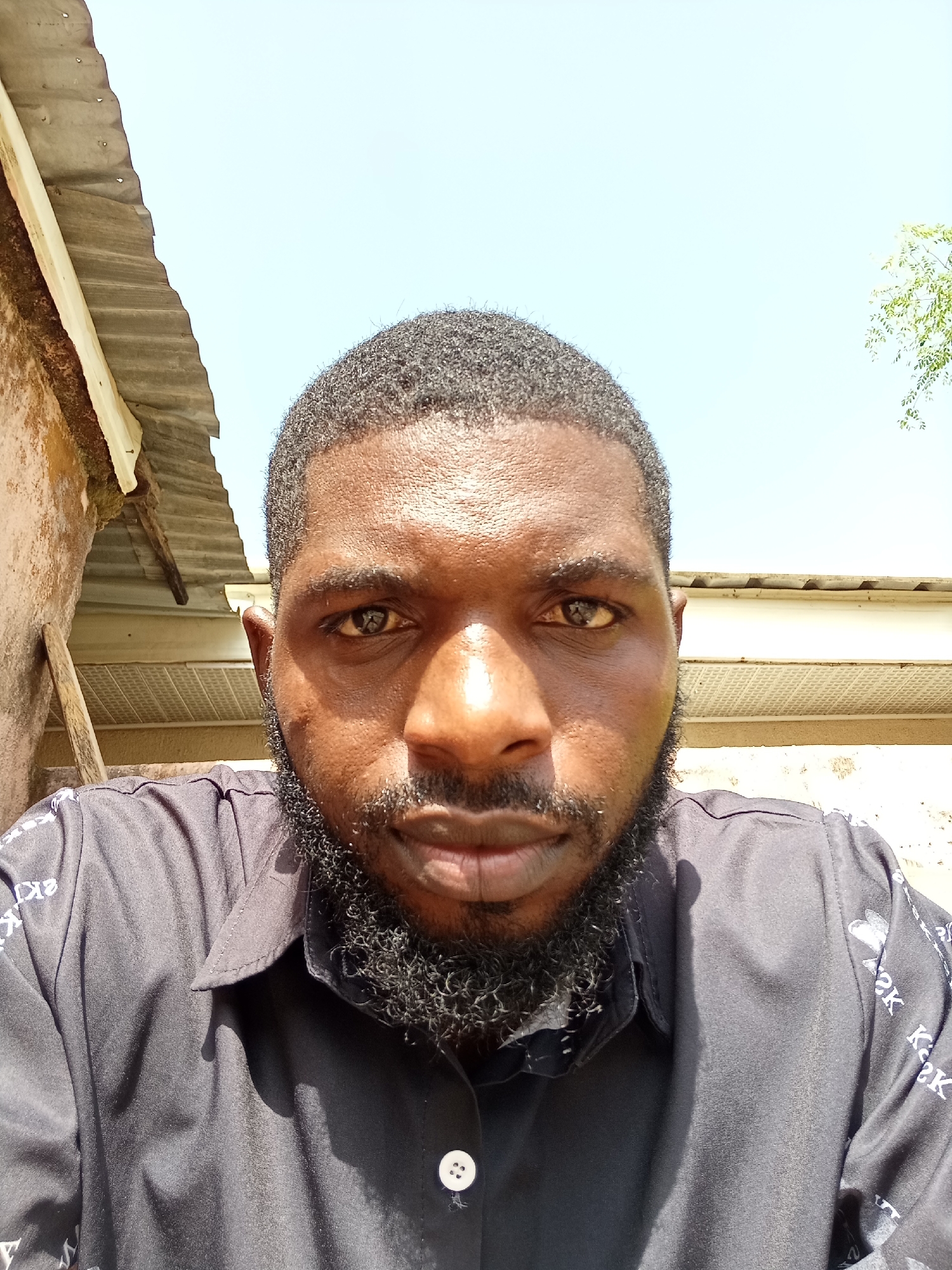 A close-up, head-and-shoulders selfie of a Black man with dark skin, a full beard, and short, curly hair is taken outdoors. He is wearing a black collared shirt with white print on the sleeves, and one white button is visible on the shirt. The man is looking directly at the camera with a neutral expression. Behind him, there is a rustic building with a corrugated metal roof and a light-colored wall. The sky is bright blue and clear, with some leaves of a tree visible in the upper right corner. The lighting suggests it is daytime. The location is identified as Rafin Par, Nigeria.