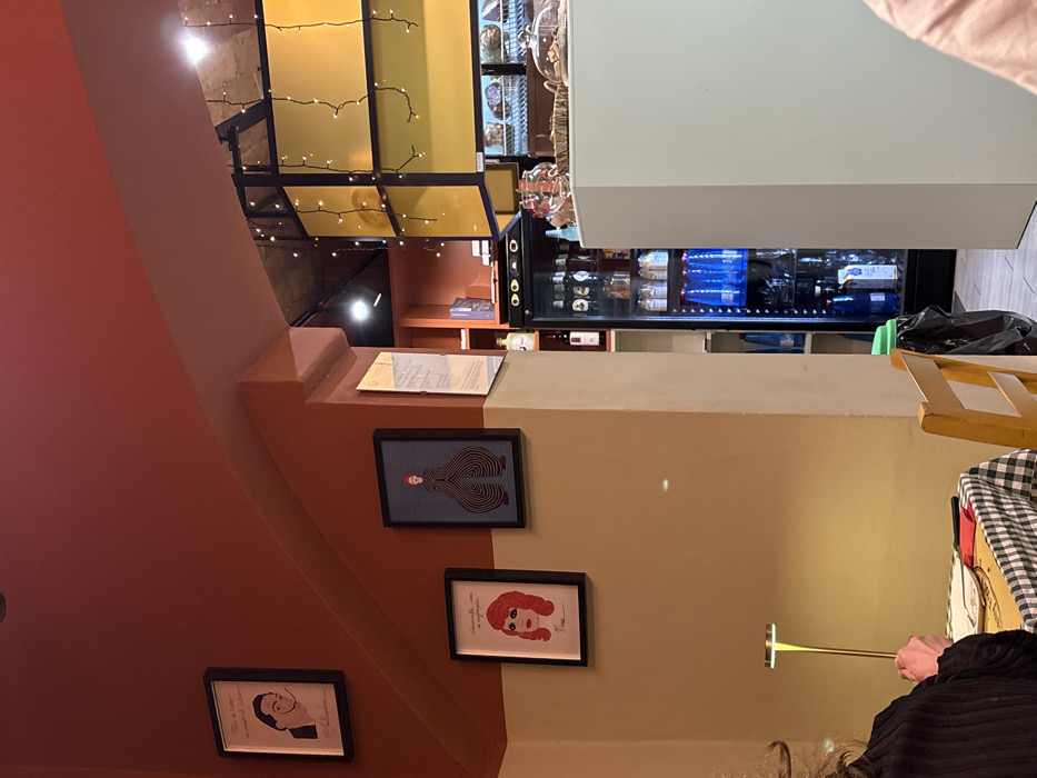 The image shows an indoor scene with an unusual perspective. A set of stairs and a wall adorned with framed pictures are visible on the left. A document rests on the stairs. Above, a decorated ceiling with lights is seen. To the right, a refrigerator stocked with blue-packaged goods and beverages stands, along with a table with a checkered cloth, and a person holding a thin golden object. The colors used are mainly brown, yellow, and white.