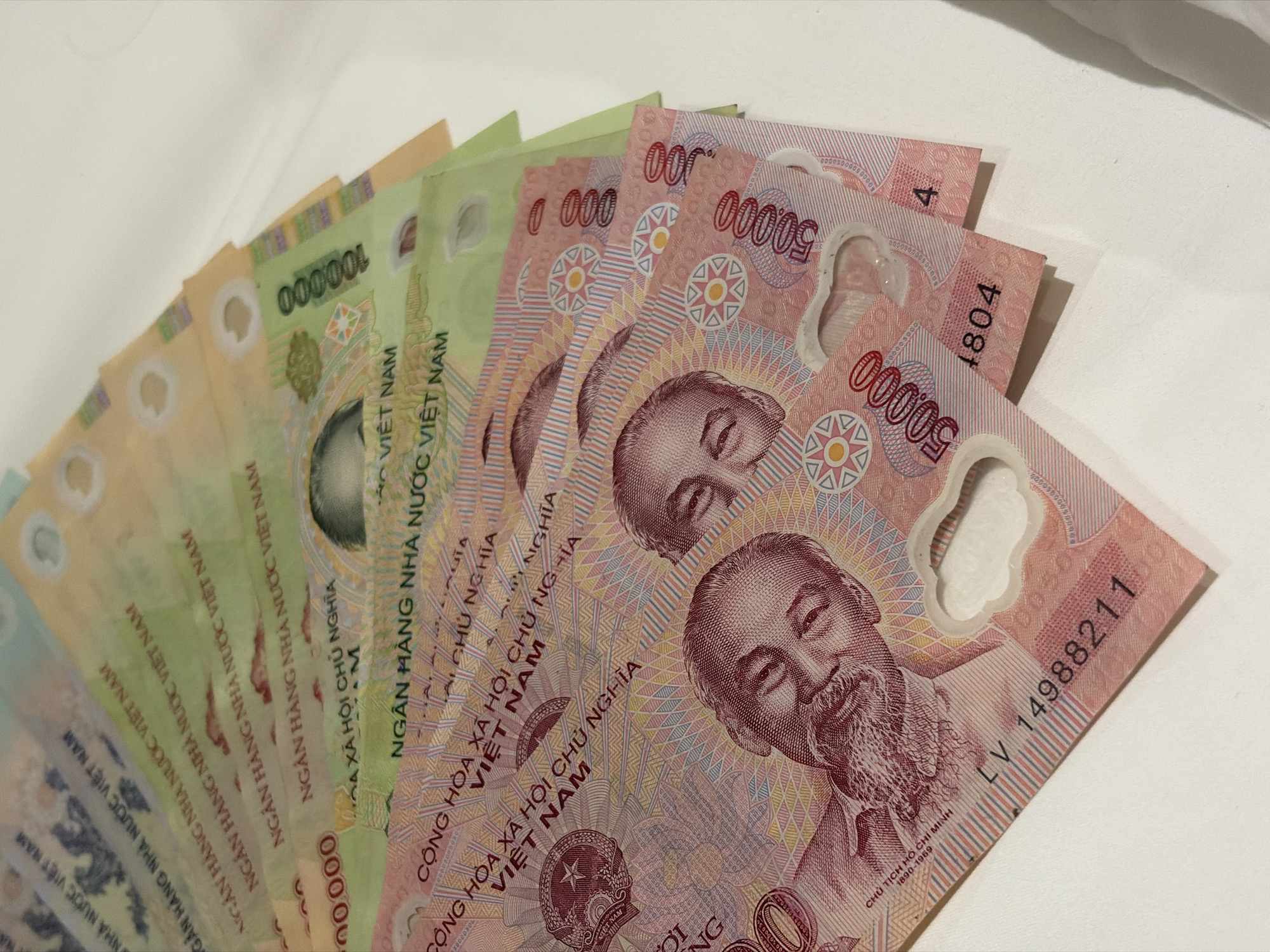 The image displays a collection of Vietnamese currency notes. The notes include denominations of 50,000 VND and 100,000 VND. All of the notes bear a portrait of Ho Chi Minh, the first president of North Vietnam. The text on the bills includes the phrases "CỘNG HÒA XÃ HỘI CHỦ NGHĨA VIỆT NAM", meaning "Socialist Republic of Vietnam," and "NGÂN HÀNG NHÀ NƯỚC VIỆT NAM," which translates to "State Bank of Vietnam." The serial number "LV 14988211" is visible on the bottom note. The photograph was likely taken indoors.
