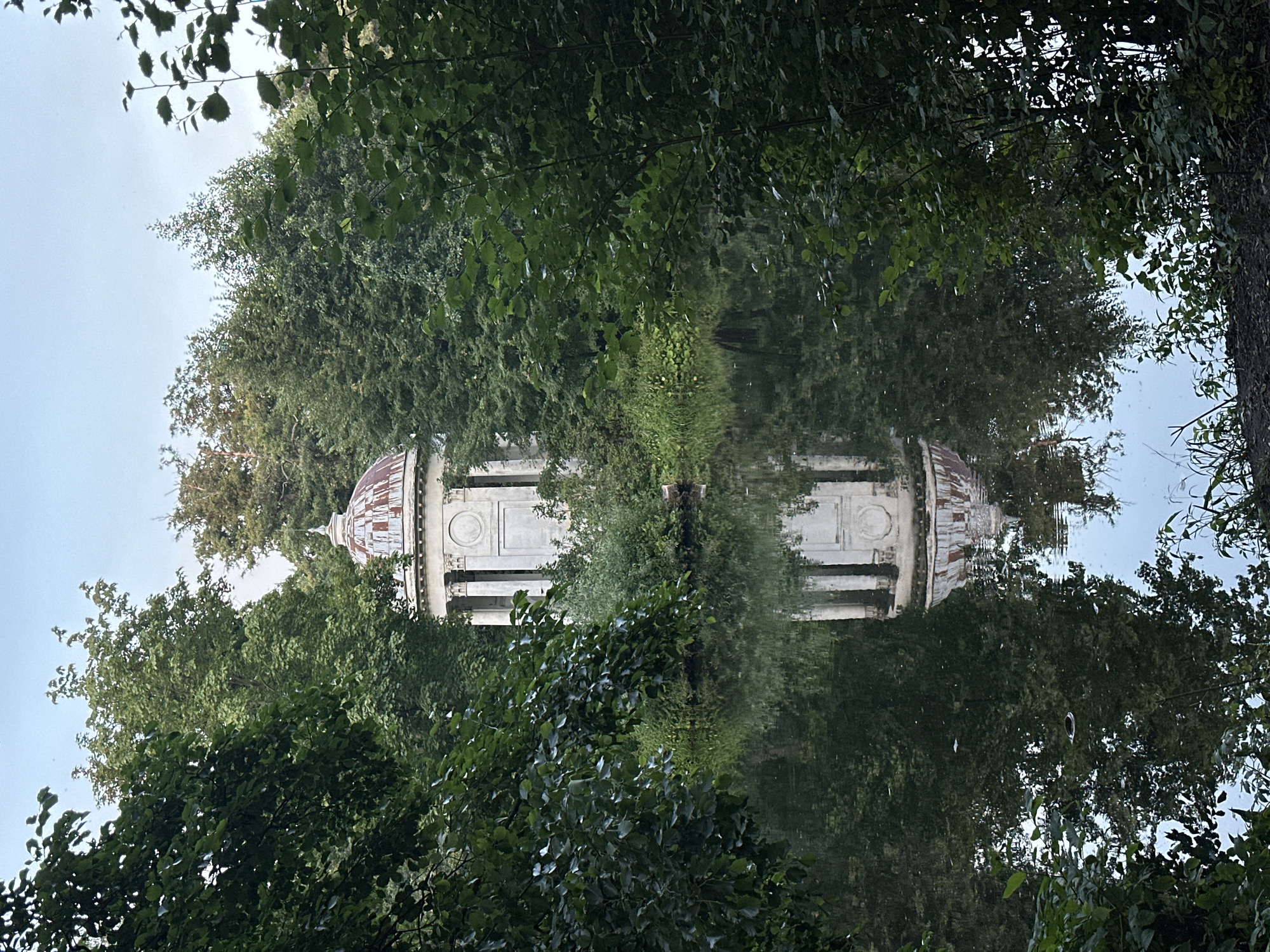 The image captures a view of the Church of Vladimir Icon of the Mother of God in Bykovo, Russia, as seen from across a body of water. The church, a white neoclassical structure with a domed roof, is partially obscured by dense green foliage that frames the foreground and midground. The water in front of the church acts as a mirror, reflecting the architectural details and the surrounding trees, creating a symmetrical composition. The sky above is a pale blue with some overcast. The overall scene is tranquil and picturesque, evoking a sense of serene nature surrounding a historical monument.