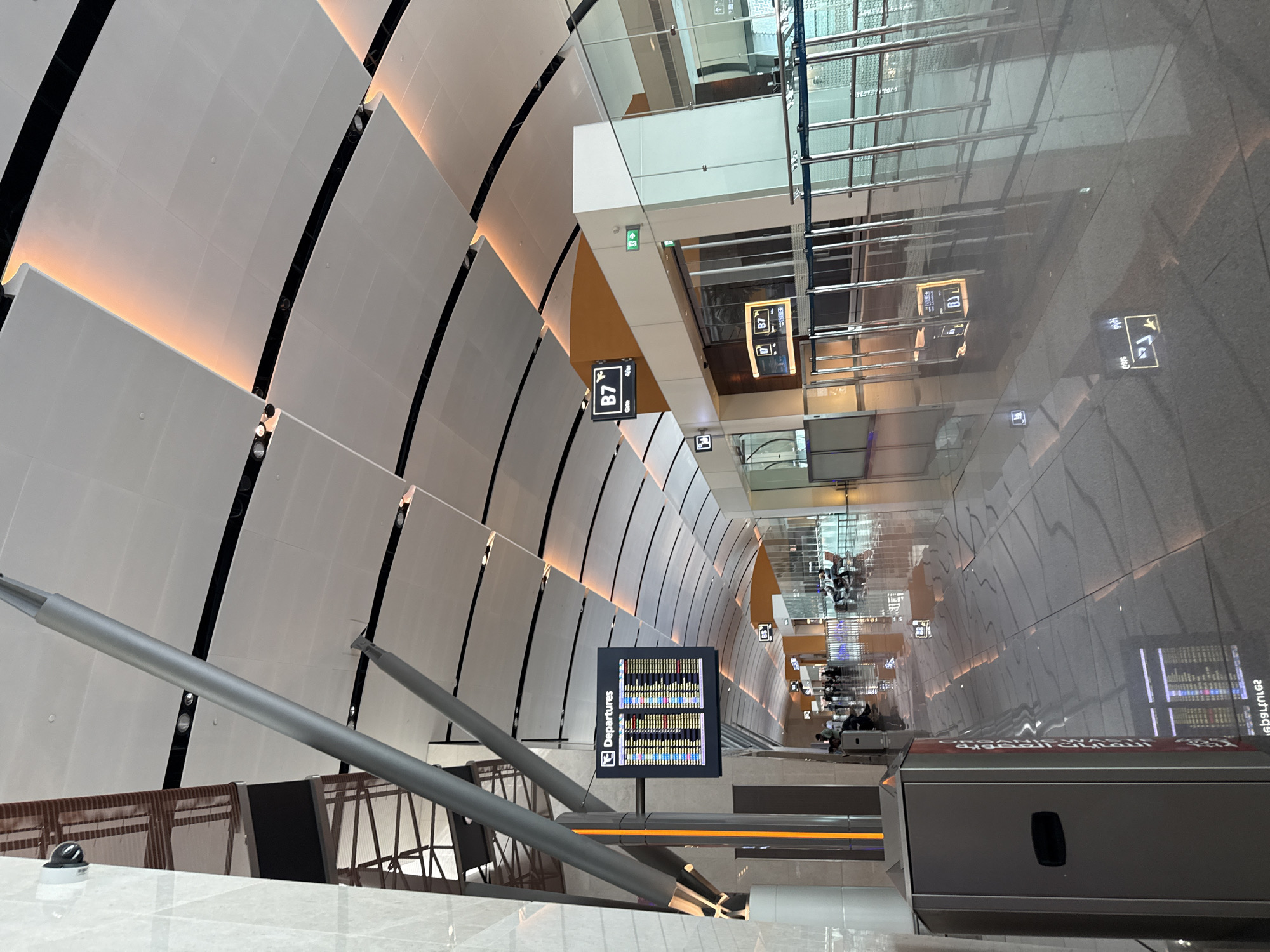 The image shows an interior view of a modern airport terminal in Seeb Province, Oman. The scene is dominated by the sleek, curved architecture of the ceiling, featuring large, white, modular panels with integrated lighting that casts a warm, ambient glow. A security camera is visible mounted on a textured, light-colored pillar on the left side of the frame.
In the foreground, a large, gray trash receptacle stands to the left. Further into the terminal, a departure board displaying flight information is mounted on a dark, rectangular screen, indicating "Departures." Two signs with the letter "B7" are also visible, one mounted higher on a wall and another on a digital display.
The floor is highly polished and reflective, mirroring the ceiling and architectural elements. Ropes and stanchions create queueing areas, and several individuals are seen sitting on benches or walking in the distance, suggesting the terminal is operational. The overall impression is one of spaciousness, modernity, and efficient travel infrastructure.