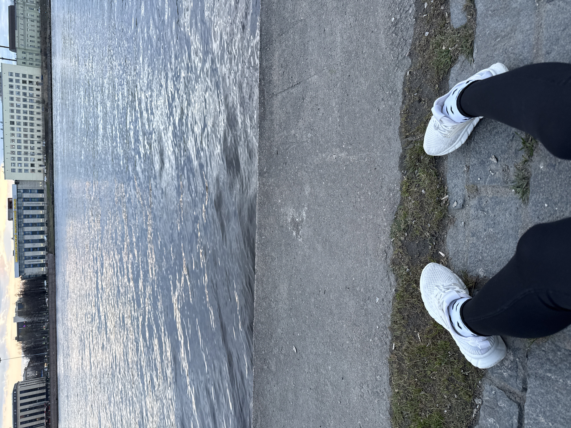 The image, rotated 90 degrees counter-clockwise, depicts a first-person view of a person's legs and feet standing by a body of water. The individual is wearing white athletic sneakers with white socks and black leggings or trousers. They are positioned on a concrete and cobblestone path, which features interspersed patches of dry grass and dirt.
To the left of the path, a wide body of water, likely a river or canal, dominates the middle ground. Its surface is rippled, reflecting the soft, diffused light of what appears to be late afternoon or early evening, with warm glints shimmering across the water.
On the opposite bank, a series of multi-story urban buildings line the waterfront. These structures exhibit a somewhat classical or utilitarian architectural style with numerous windows. On the facade of one prominent building, partial text is visible: "ИМПЕРАТОРСКИЙ" (IMPERATORSKIY), with additional letters like "СКИЙ" and possibly "АПТЕКА" (pharmacy) discernable, hinting at a historical or imperial connection. The distant skyline also shows more buildings and some sparse trees under a softly lit sky.
The scene suggests a quiet moment by an urban waterway, characteristic of cities like Saint Petersburg, Russia, aligning with the provided location context. The person appears to be paused, perhaps during a walk, observing the tranquil setting.