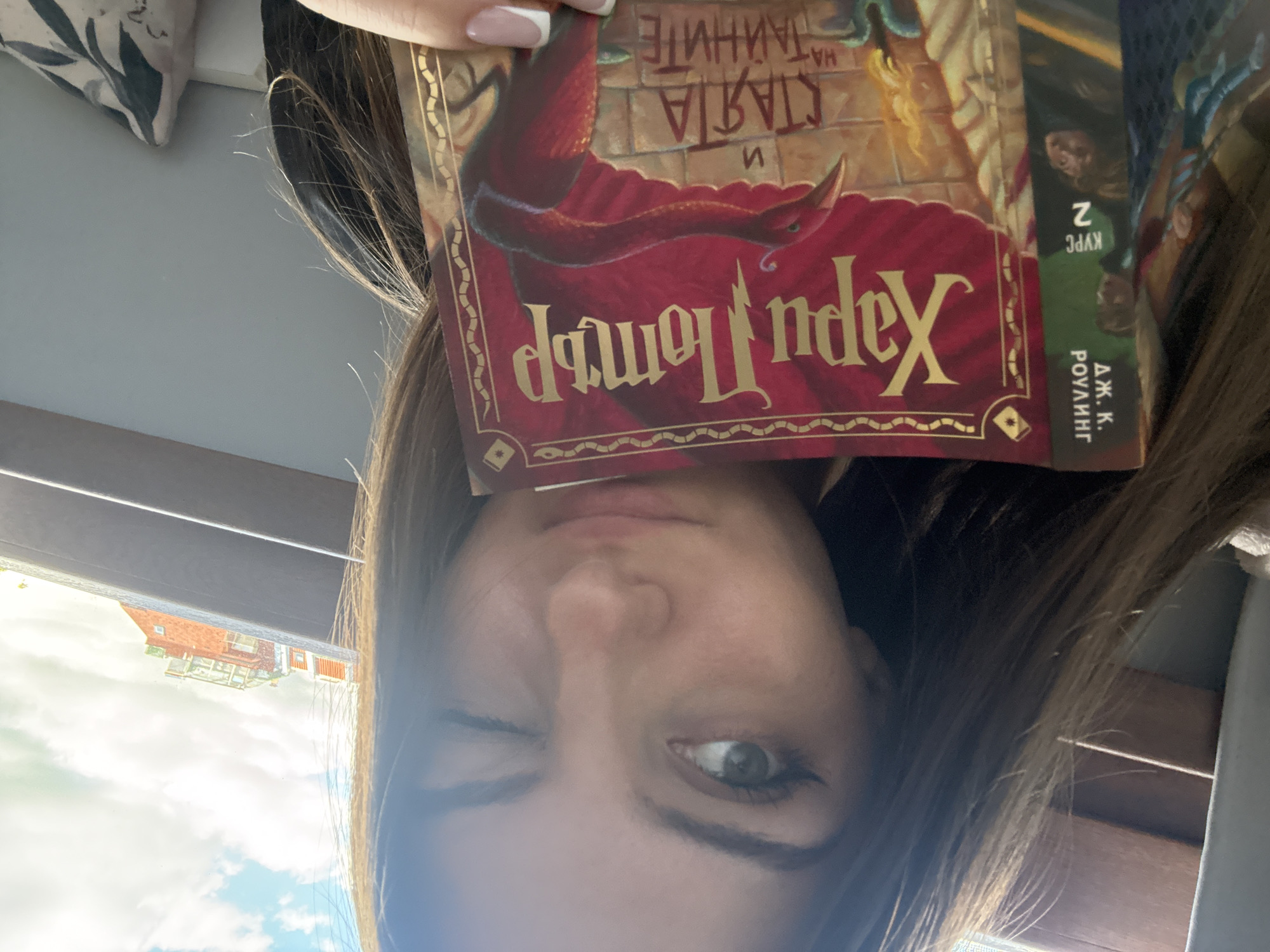 A young woman with brown hair is holding a book in front of her face. The book is titled "Harry Potter and the Chamber of Secrets" in Bulgarian ("Хари Потър и Стаята на тайните"). The woman is winking with her left eye. Through a window in the background, part of a building with a red roof is visible. The sky is partly cloudy. The image does not contain enough information to identify the location in Sofia, Bulgaria.