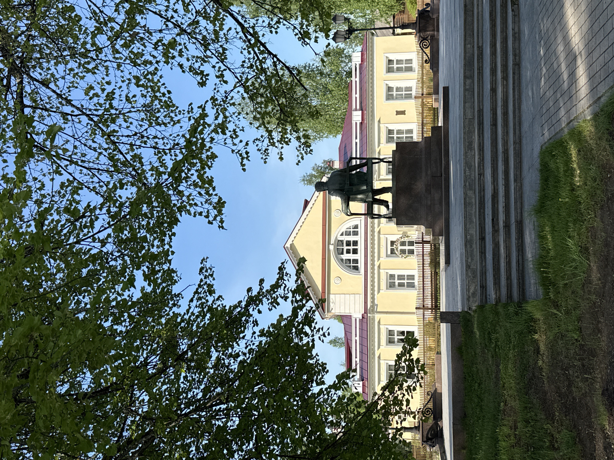 The image shows a sculpture of Pyotr Ilyich Tchaikovsky in Votkinsk, Russia. He is seated in a chair on a pedestal in front of a yellow building with a red roof. A row of stairs leads up to the sculpture. Trees with green leaves frame the scene on the left and right. The sky is blue.
