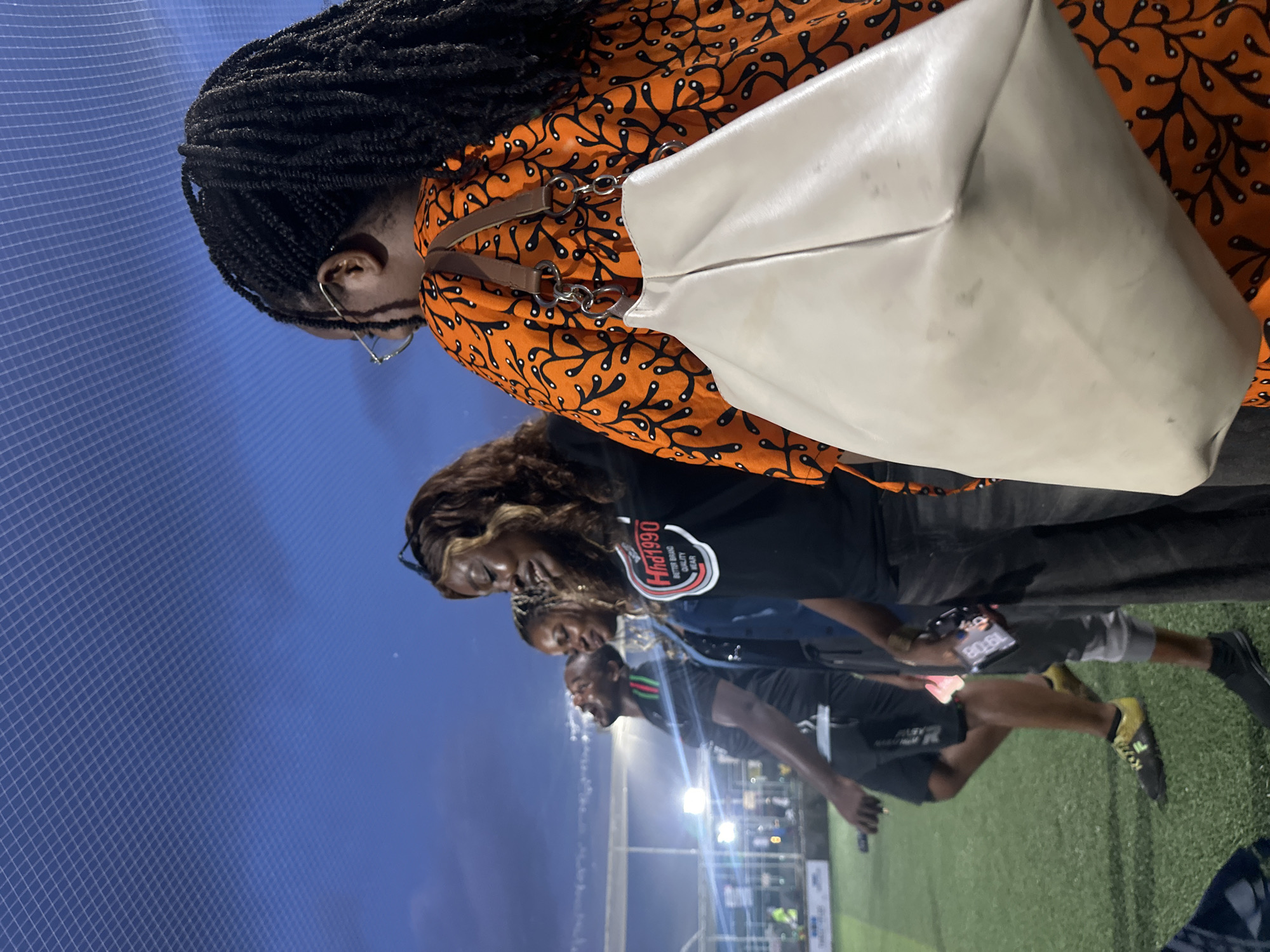 An outdoor evening photo captures four individuals on a brightly lit artificial turf field, possibly a sports pitch, under a netted ceiling. The sky is a deep twilight blue. The foreground is dominated by the back of a woman wearing an orange, black, and white patterned dress and carrying a large, light-colored bag over her shoulder. To her left, a woman with long, dark braided hair and glasses is partially visible, facing away from the camera. Next to her, a woman with long brown hair is smiling, wearing a black t-shirt with a white circular logo that reads "Hhd1990 BETTER BRAND QUALITY GEAR". To her left, a man is seen from the chest up, with his head tilted back and mouth open, wearing a black t-shirt with red and green stripes on the collar. Further to the left, another man is visible from the waist down, wearing black shorts and yellow athletic shoes. The man to his right wears gray sweatpants and black shoes. In the background, stadium lights illuminate the field, and a fence with white horizontal bars encloses the area. Several other figures are indistinctly visible in the background. The presence of an artificial turf field, stadium lighting, and what appears to be a recreational facility suggests the location could be a sports complex or stadium within Abuja, Nigeria, possibly at the National Stadium or a similar venue, given the context of outdoor sporting events.