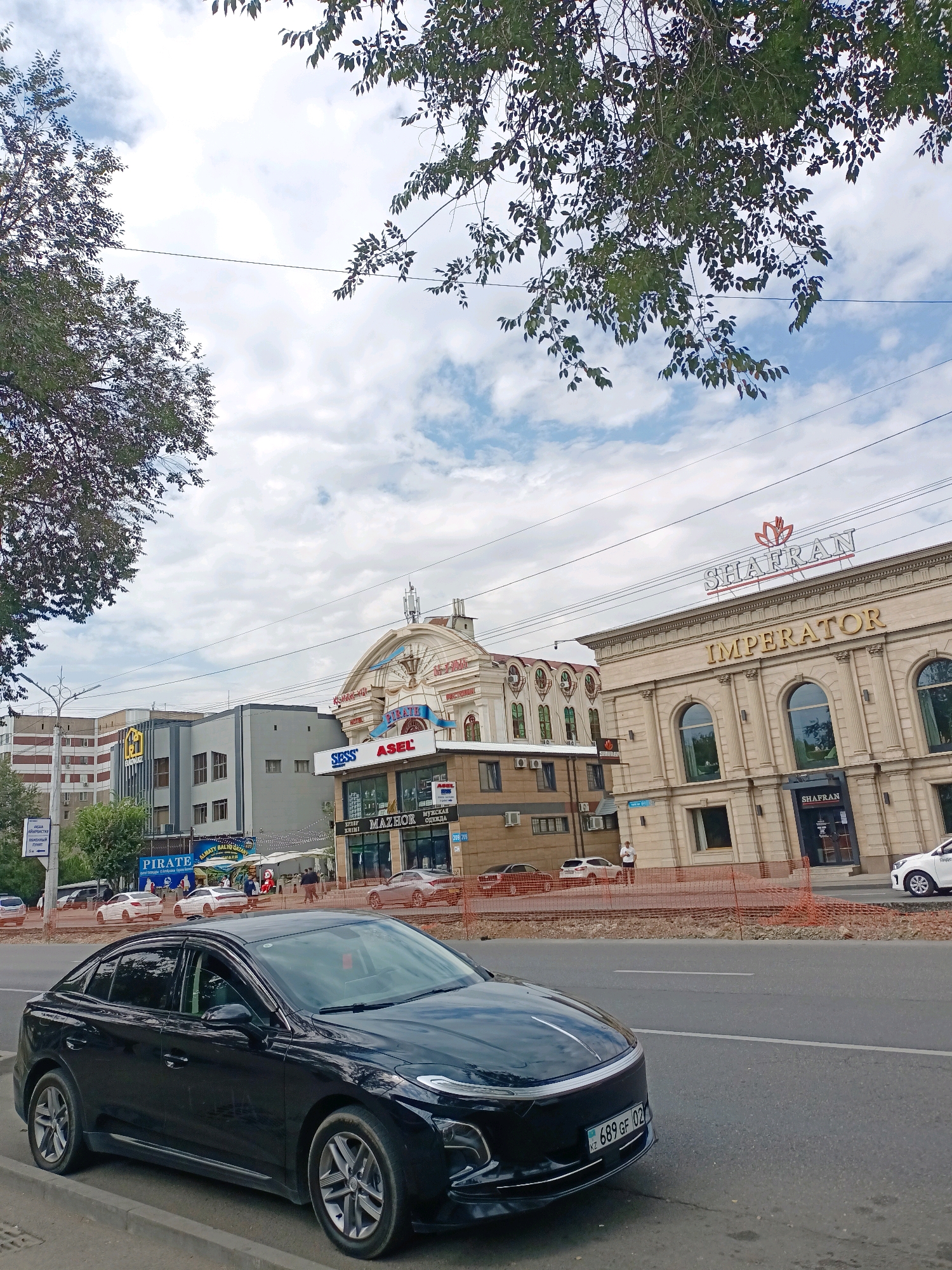 The image shows a street scene in Almaty, Kazakhstan, with a black car in the foreground parked on the side of the road. Behind the car, several buildings are visible, including one with the prominent signage "IMPERATOR" and "SHAFRAN". Another building features the sign "PIRATE". The sky is cloudy with patches of blue. There are trees with green foliage on the left and top of the frame. Utility wires are stretched across the sky. Construction barriers are in place along the sidewalk in front of some of the buildings. Several people are visible walking on the sidewalk. The license plate of the black car is visible and reads "KZ 689 GF 02".