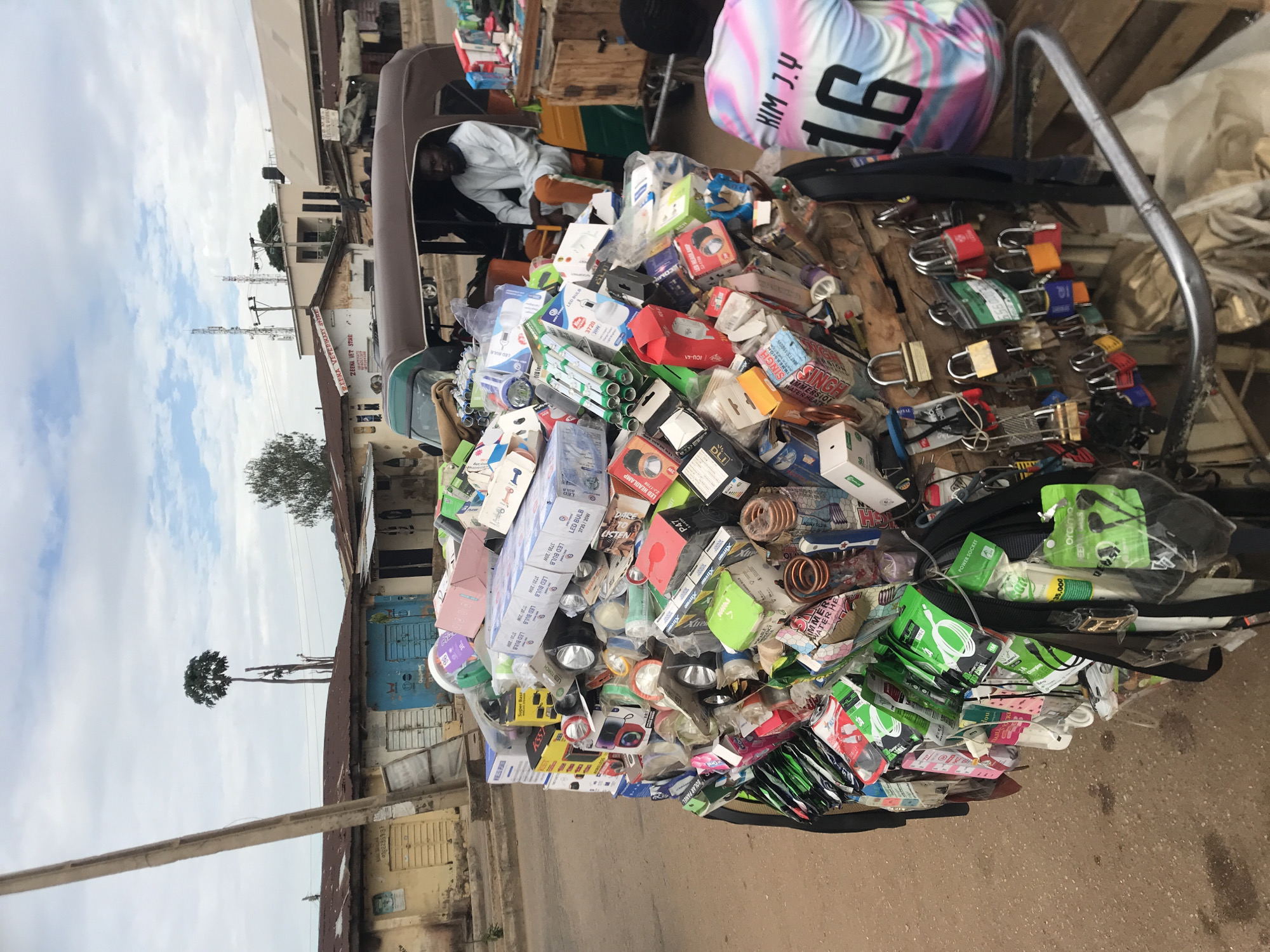 The image depicts a street scene in Rafin Par, Nigeria. In the immediate foreground, a mobile vendor's cart is heavily laden with a diverse array of new merchandise, primarily small electronics and hardware. The cart's contents include numerous boxed LED bulbs (some labeled "LED BULB 37W/20W"), flashlights, headlamps, Bluetooth speakers (one box labeled "Super Bass"), various charging cables (many in green and black packaging), earphones (one package branded "oraimo"), and immersion water heater elements. Hanging from the side of the cart are dozens of padlocks of various sizes and colors, secured by metal rings. A person wearing a multi-colored (pink, blue, white) t-shirt with "KIM J.Y" and "16" printed on the back is visible from behind, positioned to the right of the cart.
Behind the vendor's cart, a man with a beard is seated in the passenger compartment of a brown and green auto-rickshaw (tuk-tuk). He is wearing a light-colored, long-sleeved garment and orange-brown trousers. The street itself appears to be unpaved in the foreground.
In the mid-ground and background, a row of single-story buildings with corrugated metal roofs lines the street. One building on the far left has yellow doors, while an adjacent building has blue doors. Further down the street, a white building bears a red sign reading "ZEENA VET STORE." A tall, slender tree with foliage at its apex stands prominently against the sky. Utility poles and power lines are visible, with two distant communication towers rising above the rooftops. The sky is overcast with scattered clouds.