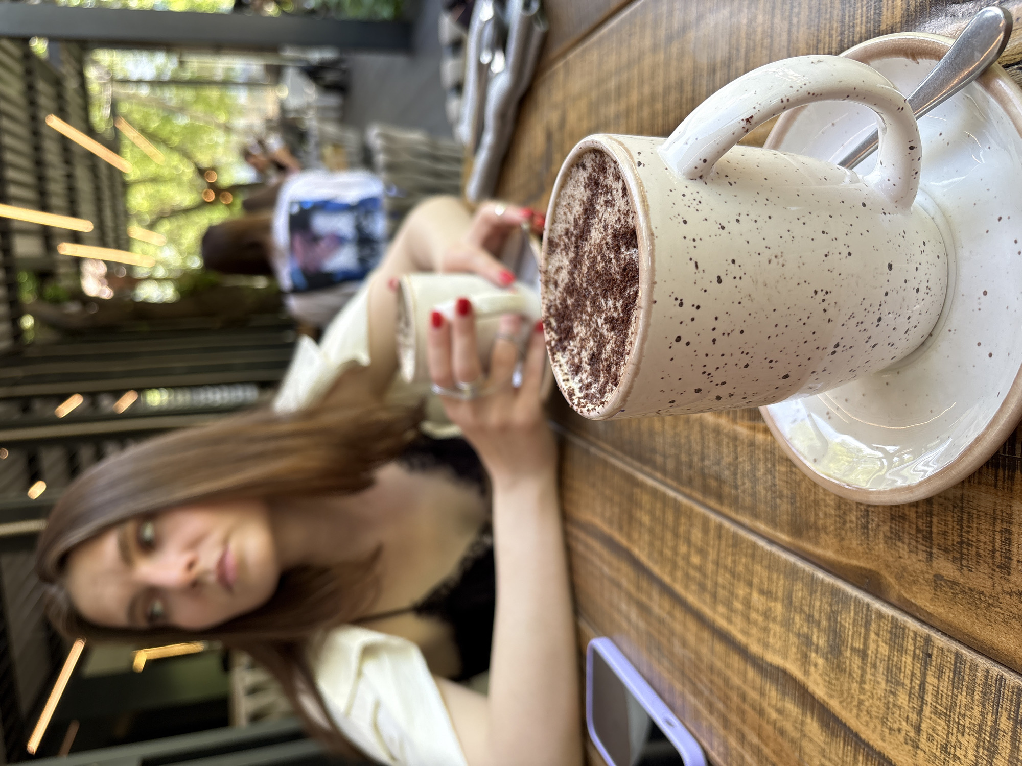 This image depicts an outdoor or semi-outdoor cafe scene in Krasnoyarsk, Russia.
In the foreground, a speckled cream-colored ceramic mug, filled with a dark beverage topped with cocoa powder or chocolate shavings, rests on a matching saucer with a silver spoon. The mug and saucer are placed on a polished wooden table composed of horizontal planks. To the left of the mug, a smartphone in a light purple case lies on the table with its screen off.
In the mid-ground, a young adult female, out of focus, is seated at the table. She has long, dark brown hair and is wearing a black lace-trimmed top under a light-colored garment draped over her shoulders. Her gaze is directed towards the left, away from the camera. Her right hand, with red painted fingernails, holds a second, similar cream-colored mug.
The background is highly blurred, revealing a bright, verdant outdoor setting with lush green foliage, suggesting trees or plants. Warm-toned, linear light fixtures are visible overhead, casting soft glows. Several other blurred patrons are seated at distant tables, including one person visible from behind wearing a white t-shirt with a blue graphic design on the back. Further details on the table to the right, such as folded napkins and cutlery, are faintly discernible.