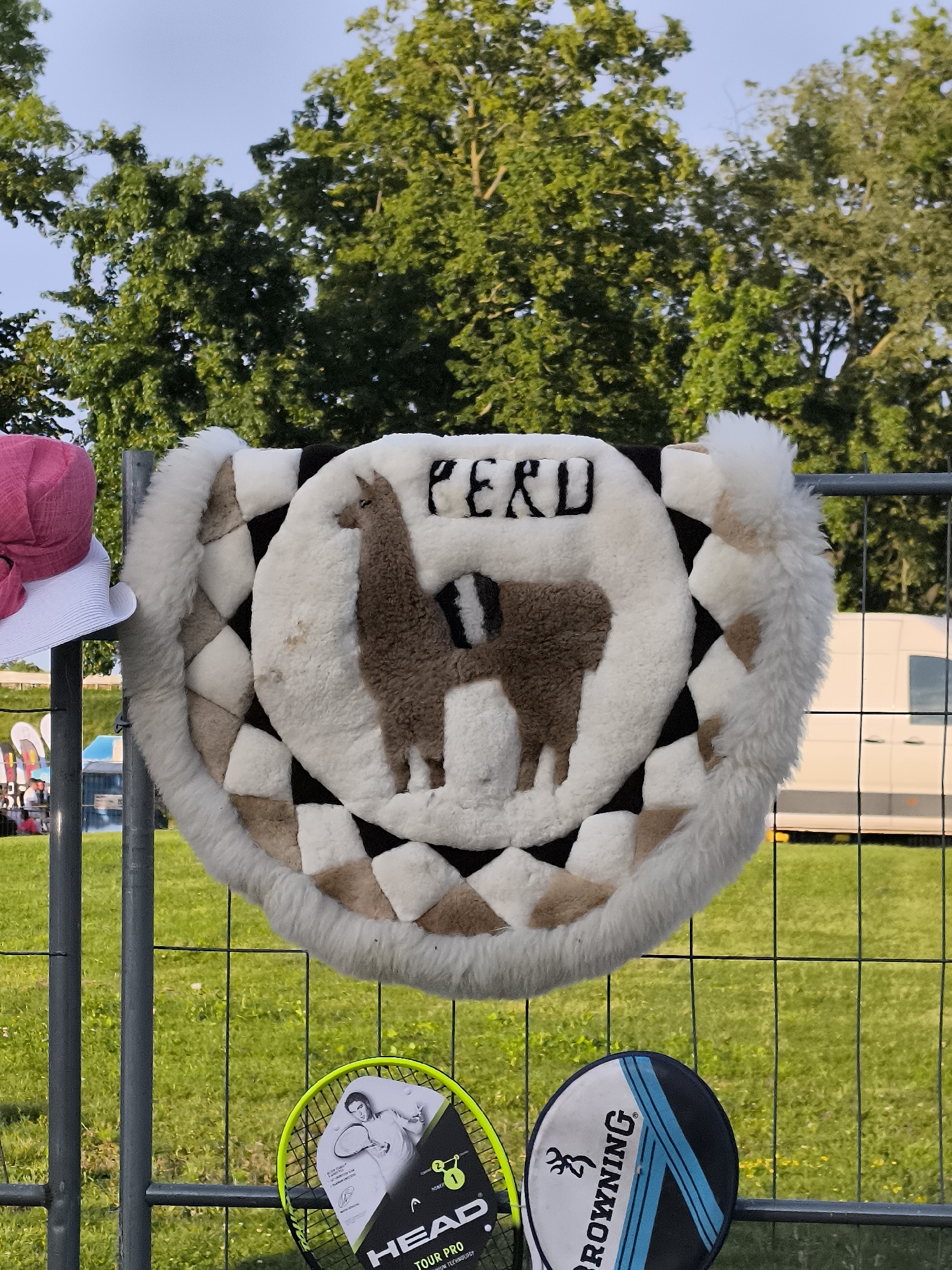 The image depicts a close-up of a decorative textile item featuring a llama and the word "PERU" embroidered on it. This item, possibly a saddle pad or wall hanging, is made of white and brown textured material with a fluffy white border. It is suspended on a metal fence. To the left of the textile, a white hat with a pink ribbon is visible. In the background, green trees, a grassy area, and a white van are seen. The lower part of the image shows parts of two tennis rackets hanging on the fence. One racket has "HEAD TOUR PRO" written on it, and the other has "BROWNING" visible. The scene appears to be outdoors, possibly at a market or festival. The provided metadata indicates the location as Klaipėda, Lithuania.