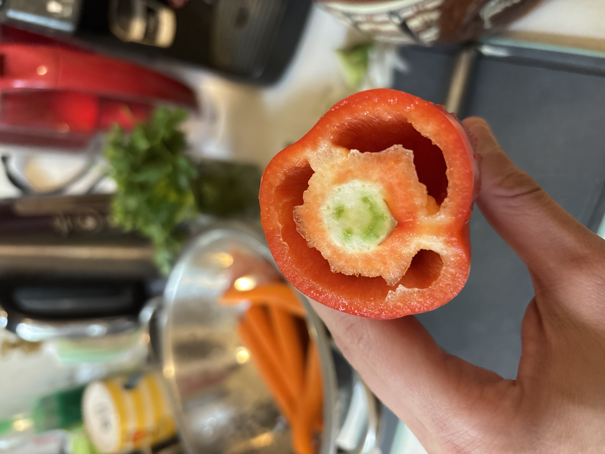 A person holds a halved red bell pepper with the cut side facing the camera. The inside of the pepper has a concentric structure with a green center. A collection of kitchen items is scattered in the background, including a red stand mixer, a bunch of parsley, a glass bowl with orange vegetables, and a bottle of seasoning. The location is not discernible from the image and thus I cannot indicate whether it is in Sofia, Bulgaria.
