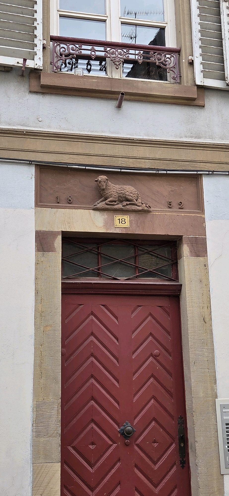 This is a medium shot, eye-level, exterior, full-frame, vertical photograph of a red wooden door and the architectural details above it. The door is centrally located and is made of several panels arranged in a chevron pattern. A ornate black metal door handle is visible on the right side of the door. Above the door, a small window with a diamond-patterned metal grate is present. Above the window is a stone inscription featuring a bas-relief of a lying sheep and numbers. The numbers "18" appear to the left of the sheep, and "52" appear to the right. A gold plaque with the number "18" is also visible below the sheep. The wall surrounding the door and window is made of stone. Further up, a brown stone ledge runs horizontally across the building. Above this ledge is a section of wall, possibly stucco, with a small metal pipe protruding from it. At the very top of the image, a white window with wooden shutters is visible. The building appears to be in Strasbourg, France, based on the architectural style. No people are visible in the image.